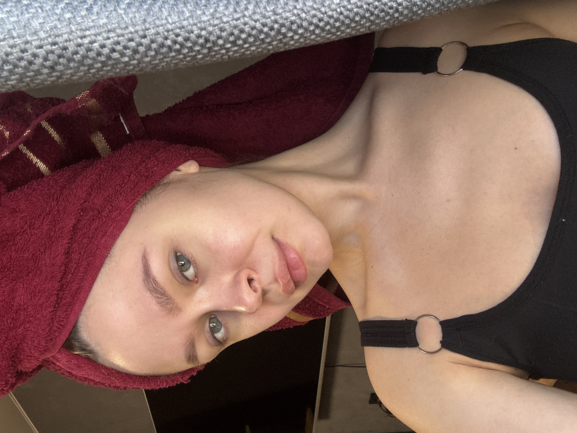 A single young adult female is depicted in a close-up portrait, facing forward with a direct gaze toward the camera. Her head is wrapped in a dark red terrycloth towel, which features visible thin gold horizontal stripes on the right side. She has fair skin, light-colored eyes, and light brown eyebrows. She is wearing a black sleeveless top or tank top, which features a silver-colored O-ring connecting the strap to the main fabric on each shoulder. The background to the left is dark and indistinct, while to the right, a light grey or blueish-grey textured fabric, possibly a blanket or curtain, is visible. The scene is an indoor setting in the city of Chaikovsky, Russia.