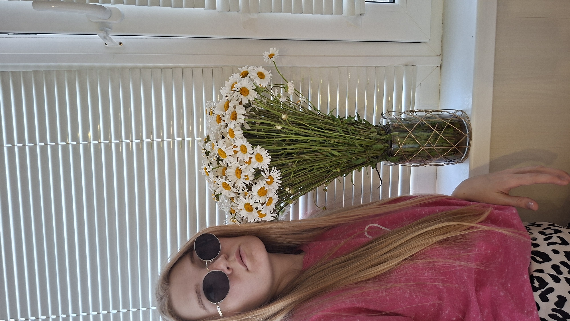 An outdoor shot taken during the day shows a young woman with long blonde hair wearing round sunglasses, a pink t-shirt, and animal-print pants. She is standing next to a large bouquet of daisies in a glass vase with a wire cage. The background consists of white horizontal blinds and a white window frame. The scene is likely in a residential area of Chaikovsky, Russia, given the presence of blinds and a window which are common in Russian apartments.