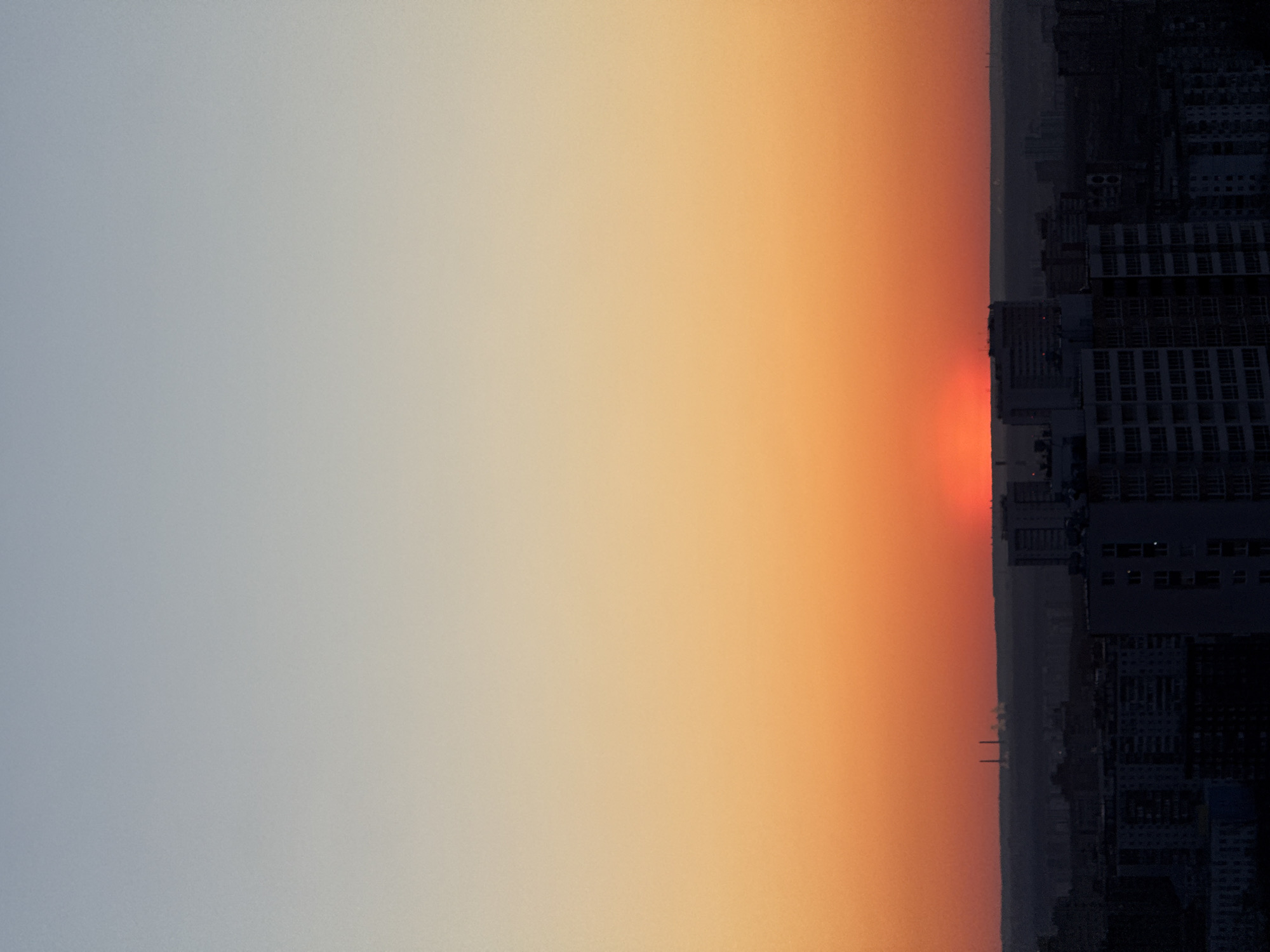 The image depicts a sunset in an urban environment. The sky transitions from light grey to orange, likely due to the setting sun. Silhouetted against the horizon is a cityscape composed of buildings, including high-rise apartments. The faint red glow of the sun is visible just above the cityscape. The buildings and the sky gradient suggest the location might be Perm, Russia.