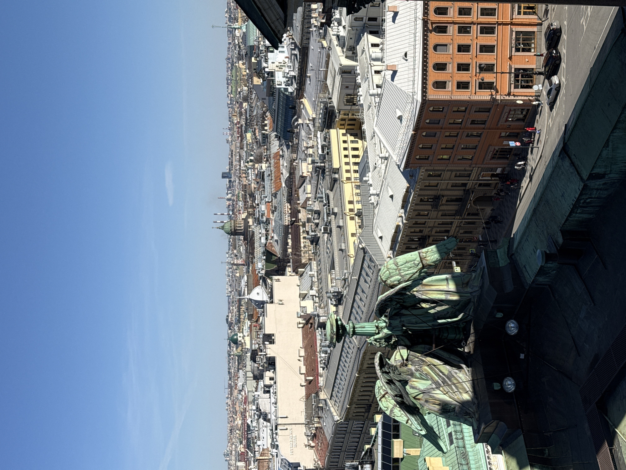 The image is an eye-level view of Saint Petersburg, Russia. The photograph appears to be taken from a high vantage point such as a tall building. The photograph depicts a cityscape with many buildings and clear blue sky. A road with vehicles, pedestrians, and traffic signs can be seen. A statue is partially in frame.