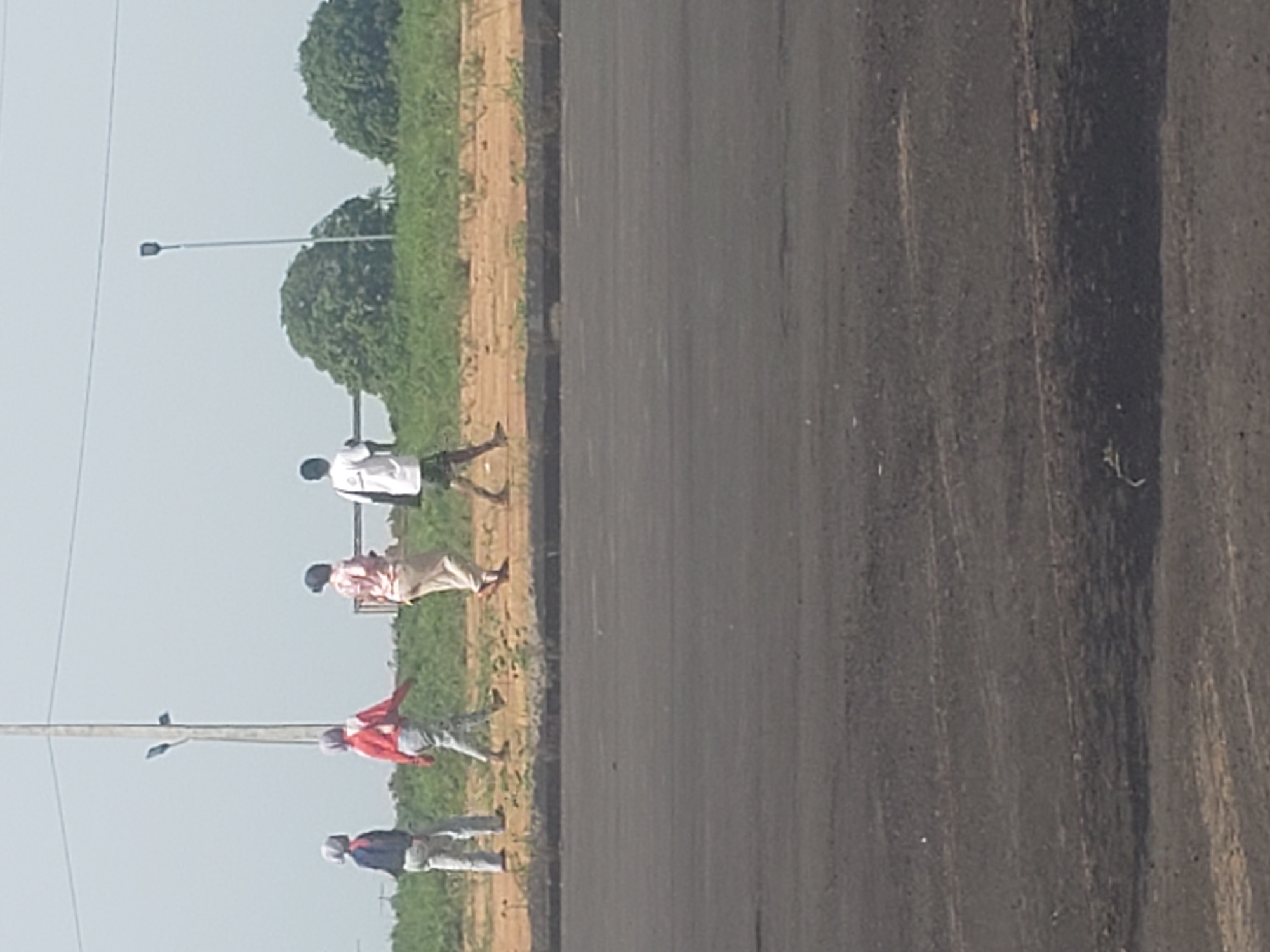 In Gumel, Nigeria, four individuals are walking on a dirt path beside a paved surface. From left to right, the first person wears a blue top and light-colored pants, with a grey head covering. The second person is dressed in a red long-sleeved shirt and light-colored pants, also with a grey head covering. The third person is wearing a patterned pink top and light-colored pants. The fourth person, a male, is in a white t-shirt and dark shorts. Behind the individuals, a grassy embankment rises, dotted with trees. A utility pole with two lights stands tall in the background, with power lines extending across the sky. The foreground is dominated by a dark, asphalt-like surface.
