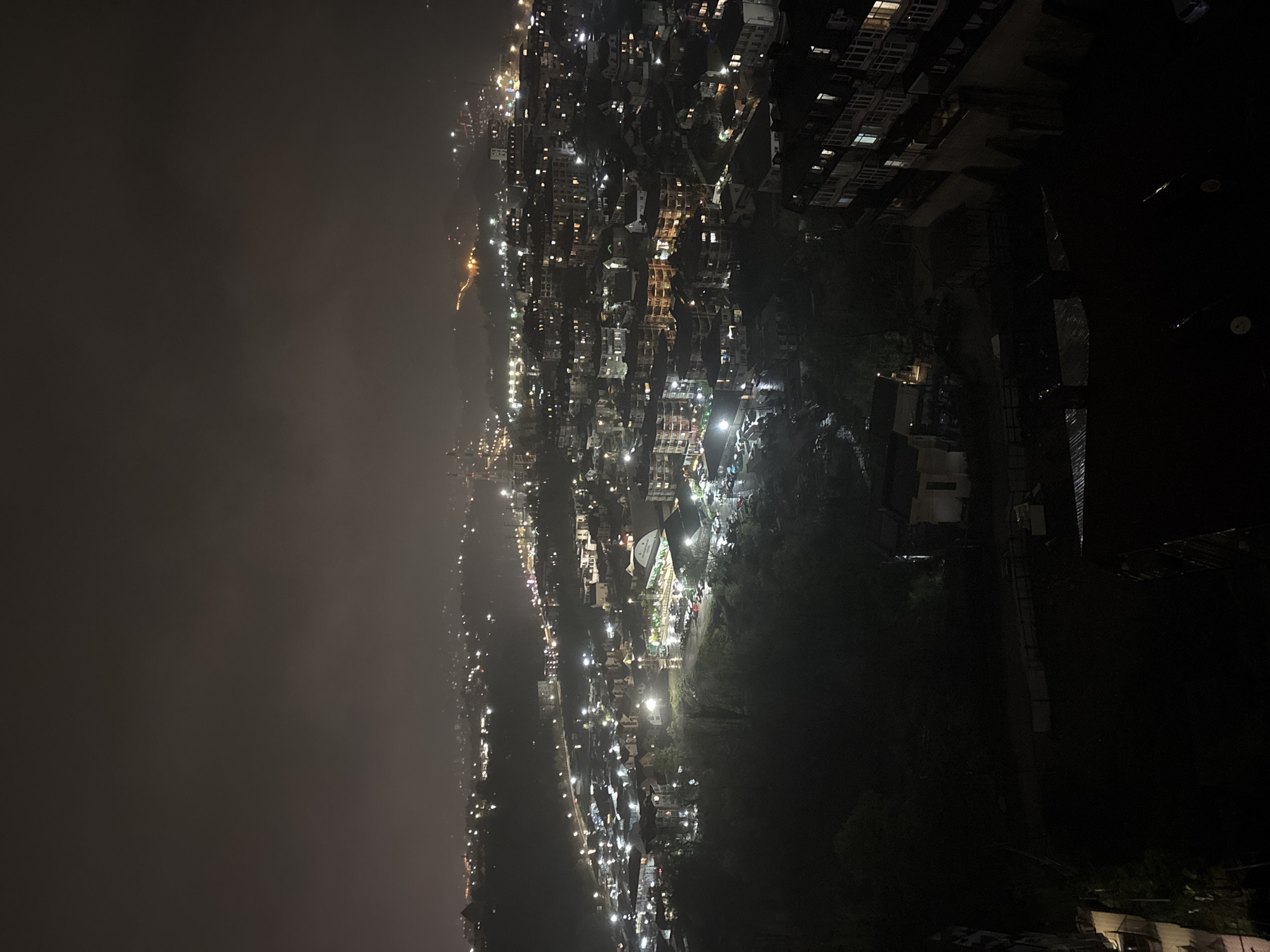 An elevated night view of a city, possibly in Sochi, Russia, is filled with buildings, lights, and haze. The city stretches across the frame from right to left, illuminated by streetlights, building lights, and vehicle headlights. On the left, a heavy fog or haze obscures the distant landscape. The right side of the image depicts dense urban development with buildings and pockets of greenery. Tall buildings are partially visible in the top-right corner. The perspective suggests the photo was taken from a high vantage point, overlooking the city.