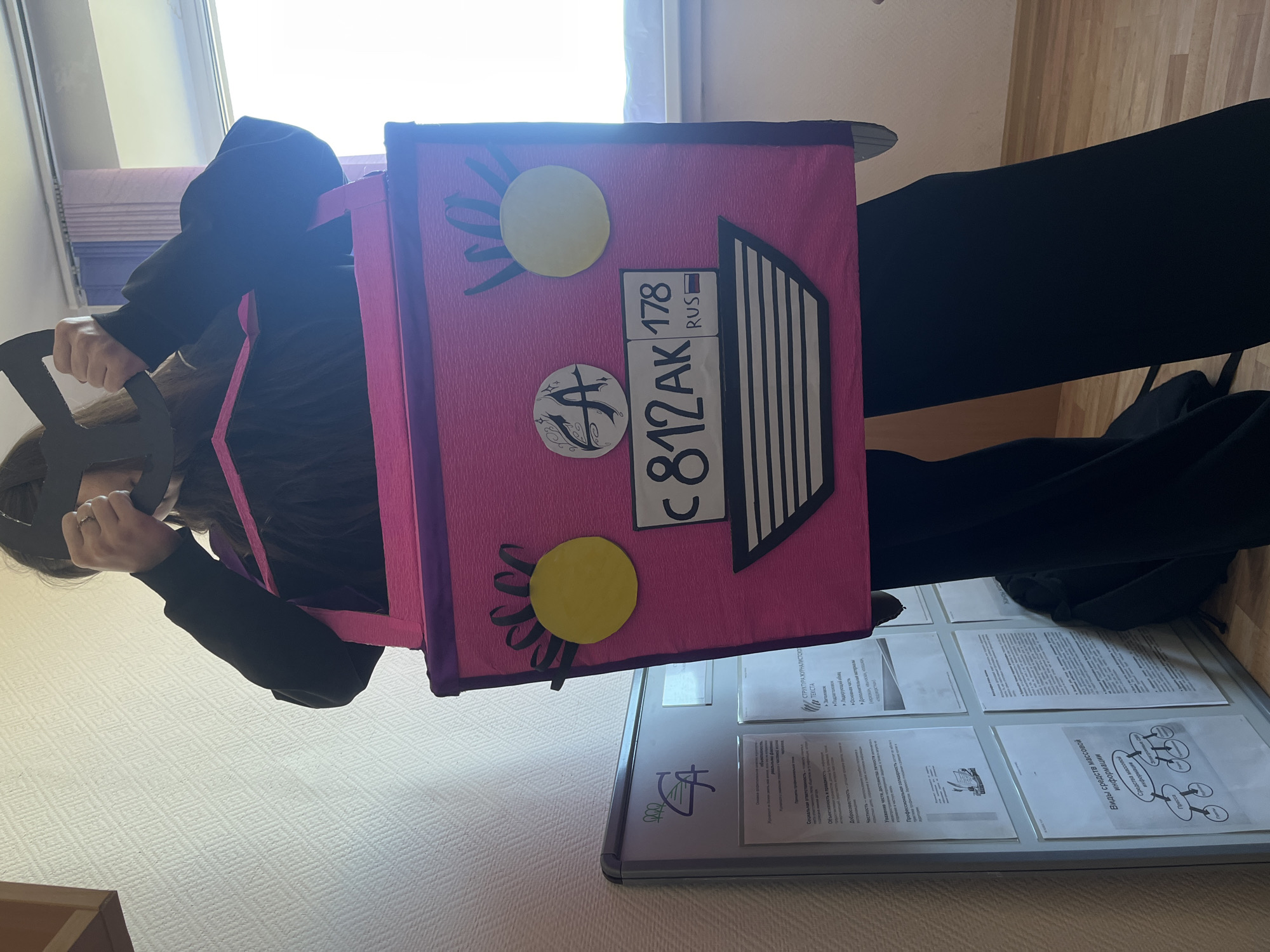 The image depicts a person wearing a black outfit, holding a pink box with Russian license plate elements (C812AK 178 RUS). The box has decorative elements like yellow circles representing the sun. The person appears to be in an indoor setting, likely a school or office, with a bulletin board behind them that contains text and diagrams. Based on the Russian license plate, the image is likely taken in Russia. Given the license plate number, it is likely to be in Saint Petersburg.