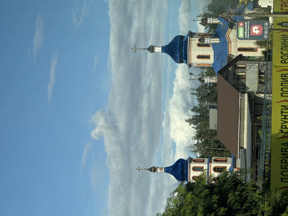 The image depicts an outdoor daytime scene on Peremohy Street, Ukraine, under a bright blue sky partially obscured by white and grey cumulus clouds. The primary subject is an Orthodox church complex featuring multiple domed structures. Two prominent cupolas are visible, both topped with reflective blue domes and gold-colored Orthodox crosses. The main body of the church is constructed from light-colored brick or stucco, accented with reddish-orange trim around its arched windows and cornices. Several smaller blue domes with crosses are also present towards the right and rear of the main structure.
In front of the church, slightly to the left, stands a building with a brown, steeply pitched roof and dark wooden siding, obscuring the lower section of the church. Additional wooden structures are visible adjacent to it. A line of coniferous trees is present in the background behind the buildings.
The lower foreground is dominated by a large yellow banner or sign displaying Ukrainian text, partially legible as "ДОБРИВА ГРУНТИ ПОЛИВ РОСЛИНИ" (Fertilizers, Soil, Irrigation, Plants), indicating an agricultural or garden supply business. To the far right, a vertical pole supports a rectangular sign featuring a green "ФОРА" (Fora) logo. White text on this sign indicates operating hours "з 6:00 до 23:00" (from 6:00 to 23:00) and an address: "м. Київ, вул. Кудряшова, 44Б" (Kyiv, Kudryashova Street, 44B). No individuals are visible within the frame. The scene is well-lit by natural daylight.