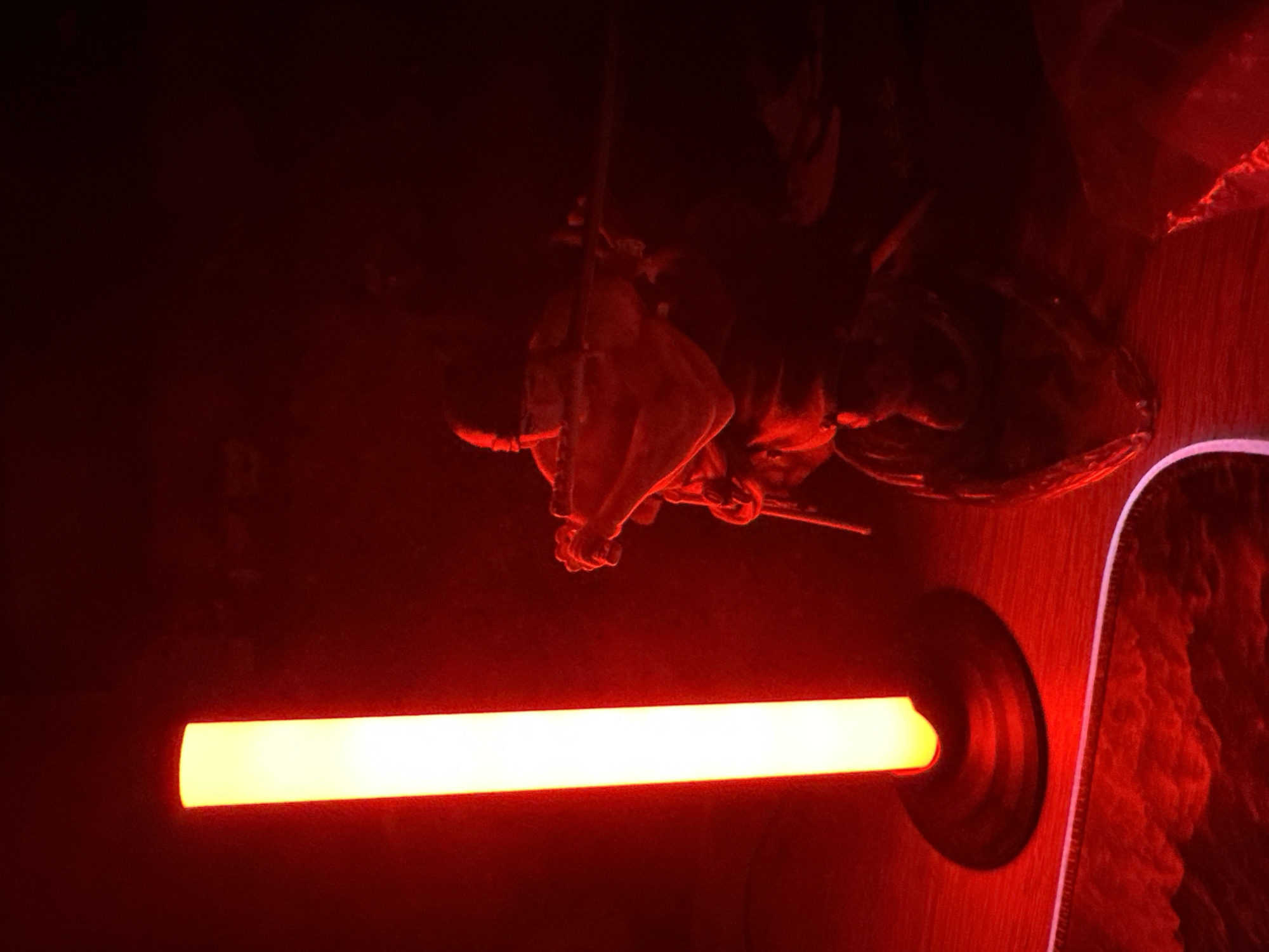 The scene depicts a dark interior space illuminated primarily by red light. On the left, a vertical, cylindrical lamp emits a bright yellow-orange light, casting a stark contrast against the predominantly red environment. This lamp has a dark, circular base. To the right, a dark humanoid figure, likely a statue or action figure, is visible, heavily silhouetted and illuminated by the red light. The figure appears to be a warrior, wearing a head covering and dark attire, holding a long, slender staff or weapon horizontally with both hands, positioned in a dynamic stance. The surface beneath the lamp and figure, presumed to be a tabletop, reflects the red light. In the bottom left foreground, a section of a dark mat or mousepad with a textured, wave-like pattern is visible, also reflecting the red ambient light. The background is indistinct and shrouded in darkness.