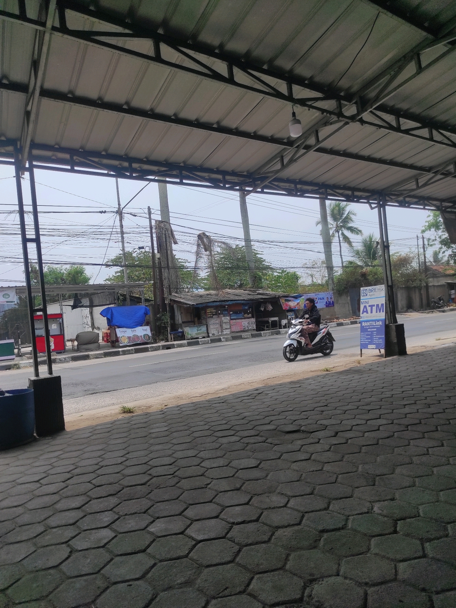 In Cipatat, Indonesia, a person rides a white scooter along a paved road. The scooter rider, wearing a dark jacket and patterned pants, is positioned on the right side of the road. The environment is characterized by a covered outdoor area with a metal roof supported by dark metal beams, above a sidewalk paved with hexagonal concrete blocks. Beyond the road, several small buildings and stalls are visible, some with corrugated metal roofs, and a blue tarp covers an object. Overhead, a dense network of electrical wires crisscrosses the sky. A "BRI Link Mini ATM" sign stands on the right, near a pole. Palm trees are visible in the background, suggesting a tropical climate. The overall scene depicts a street view with commercial activity.