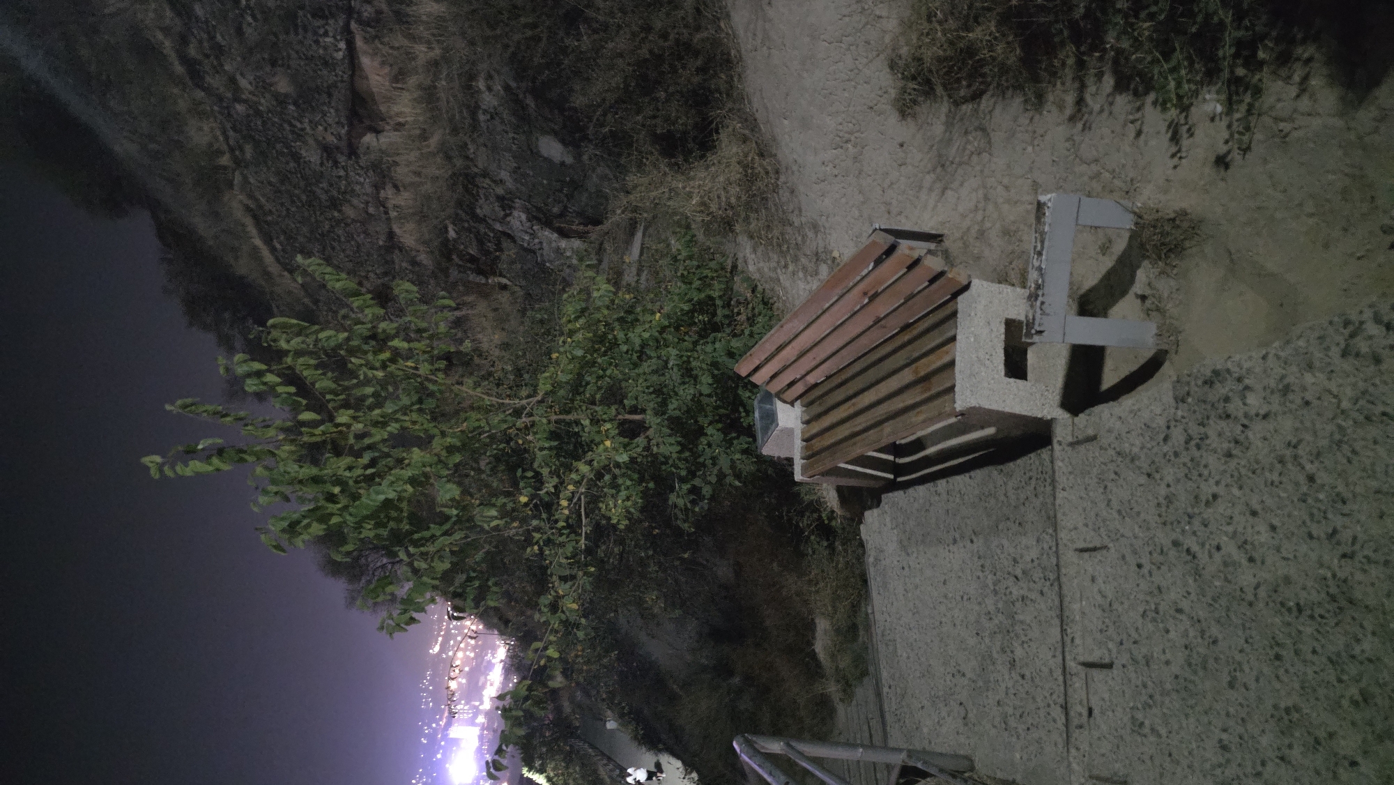 A nighttime view captures a concrete staircase leading down a rocky hillside. A wooden bench with a concrete base is positioned on a sandy area beside the stairs. In the background, a tree with green leaves stands prominently, and beyond that, a city skyline glows with lights against the dark sky. The urban landscape of Osh City, Kyrgyzstan, is visible in the distance.