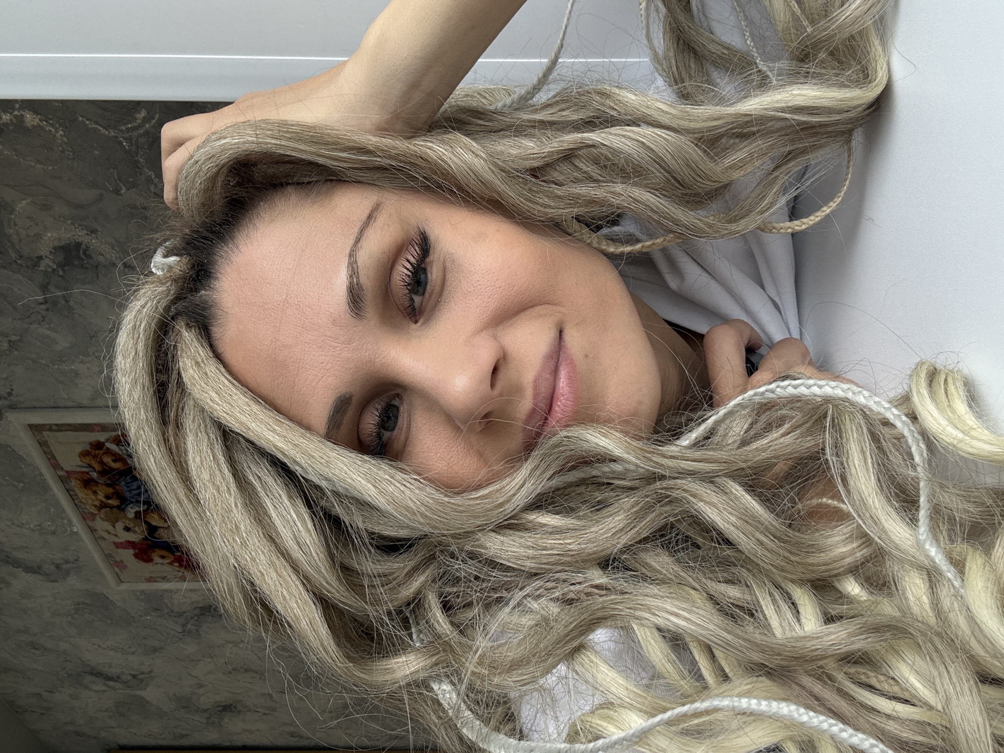 A young woman with blond hair, styled with braids and waves, is centered in the frame. Her face is visible, she is looking toward the camera with a slight smile. She is wearing dark eye makeup. Her right hand is touching her head, near her forehead. She appears to be indoors, possibly in a residential setting, as a portion of a wall and artwork are visible in the background. The background shows the inside of a building. The exact location in Rusanovo, Russia cannot be determined from this image.