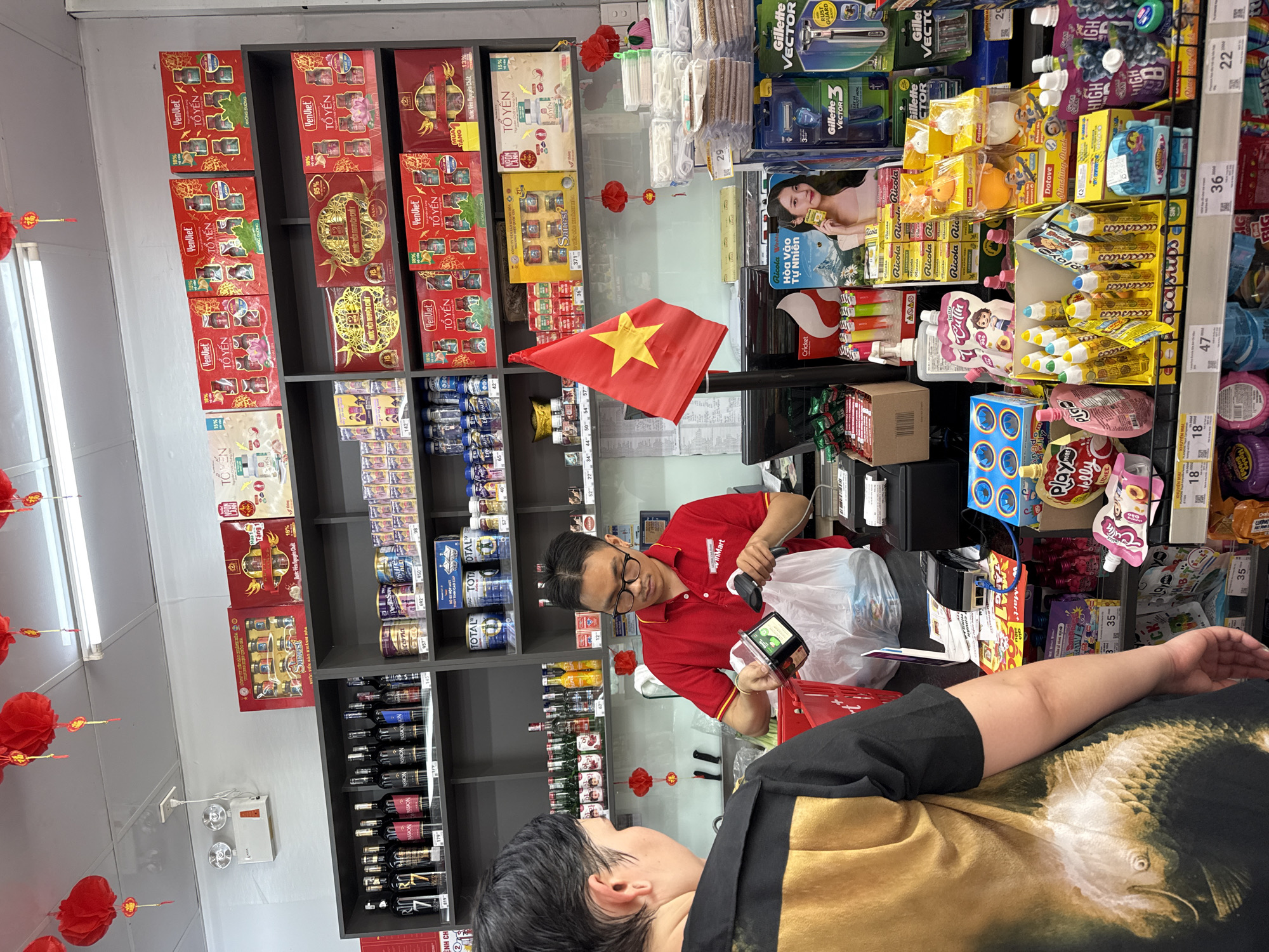 In a retail environment identified as Bac Nha Trang Ward, Vietnam, a cashier in a red uniform scans a product for a customer. The cashier, wearing glasses and a name tag that reads "VinMart," holds a barcode scanner to a black and clear plastic container. The customer, facing away from the camera, wears a black shirt with a large, detailed print of a fish. The background reveals shelves stocked with various goods, including beverages, packaged foods, and personal care items. Red lanterns are suspended from the ceiling, indicating a festive or cultural occasion. A Vietnamese flag is prominently displayed on a pole. The scene captures a typical transaction in a Vietnamese convenience store.