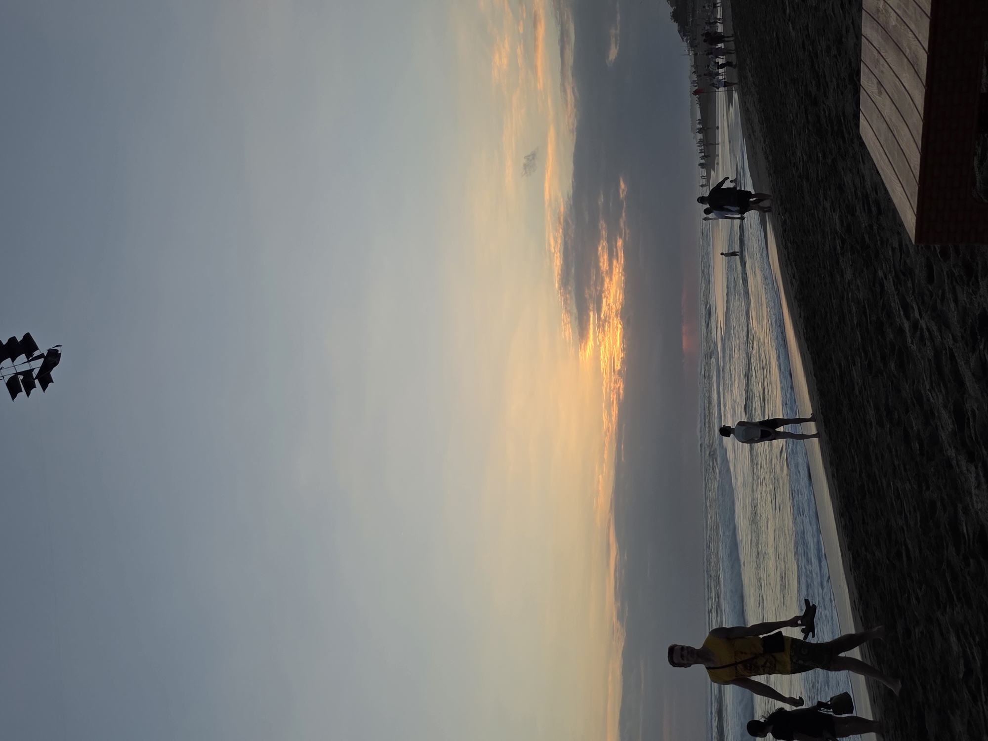 The image depicts a beach scene at dusk in North Kuta, Indonesia. The sky is filled with soft, muted colors as the sun sets, casting a warm glow on the horizon. Waves gently roll onto the shore of a dark sand beach. Several people are visible, some walking along the water's edge, while others are further down the beach. A man in a yellow tank top and patterned shorts is in the foreground, walking towards the left side of the frame, holding a pair of sandals. To his left, a person in a dark outfit carries a bag. In the background, other individuals and groups are scattered along the beach, some appearing to be wading in the ocean. The overall atmosphere is peaceful and serene, characteristic of a beach at the end of the day.