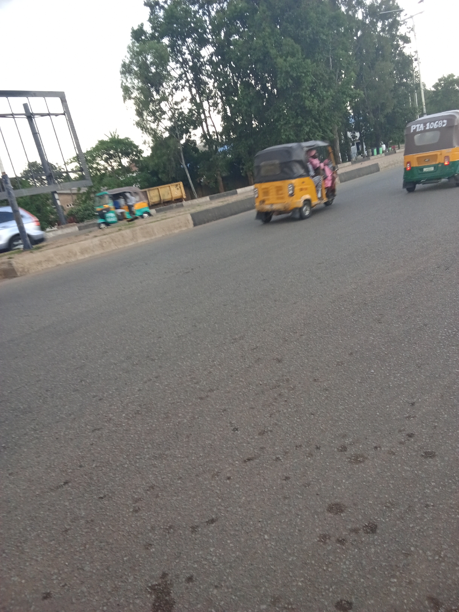 The image captures a scene on a street in Jos, Nigeria, likely during the day with overcast skies. The foreground is dominated by a grey asphalt road. In the mid-ground, there are three auto-rickshaws, also known as Keke Napep. One auto-rickshaw, yellow with a black roof, is moving away from the viewer. Another auto-rickshaw, green and yellow, is also moving away and has the license plate "PTA 10682" visible. A third auto-rickshaw, green and yellow, is parked on the side of the road. On the left side of the road, a silver car is partially visible. In the background, lush green trees line the street, and a large, dark metal structure, possibly a billboard frame, is visible. The environment appears to be urban, with a paved road and surrounding vegetation.