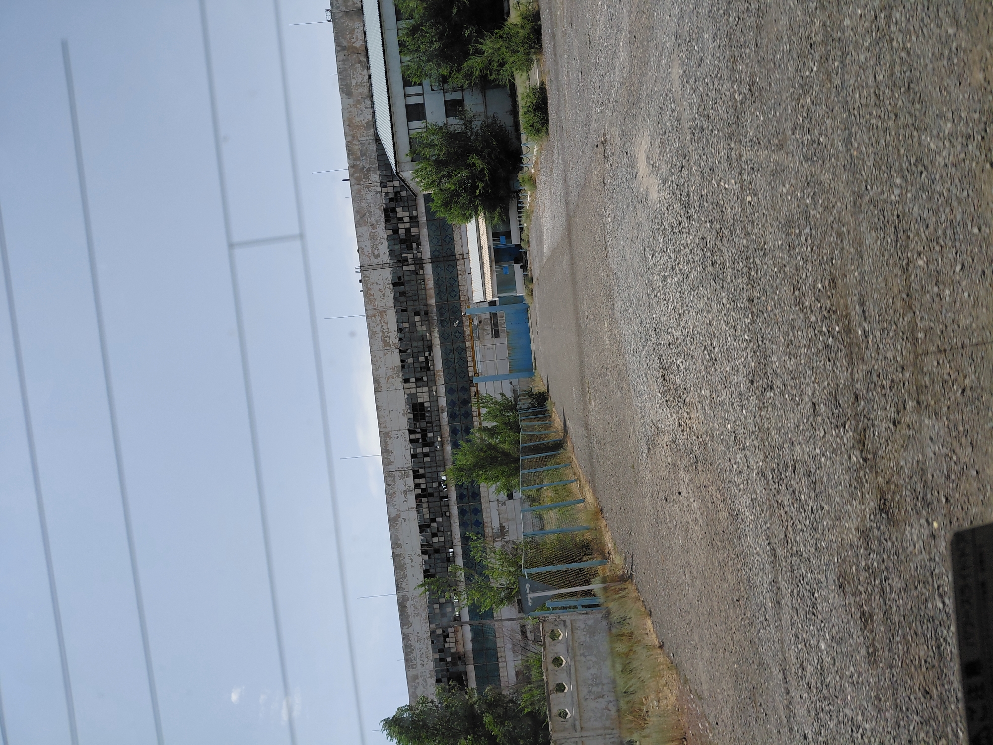 The image shows a long, weathered industrial building with many broken windows, suggesting it is abandoned or derelict. In front of the building, there is a gravel road leading towards a blue gate and a smaller building. A chain-link fence with a light blue top runs along the left side of the road, separating it from a low concrete wall. Trees and dry grass are visible on the left side of the frame. The sky is cloudy and overcast. The overall scene appears to be in a state of disrepair, possibly within an industrial area of Ақтүбек, Kazakhstan. There are no people or animals visible in the image.