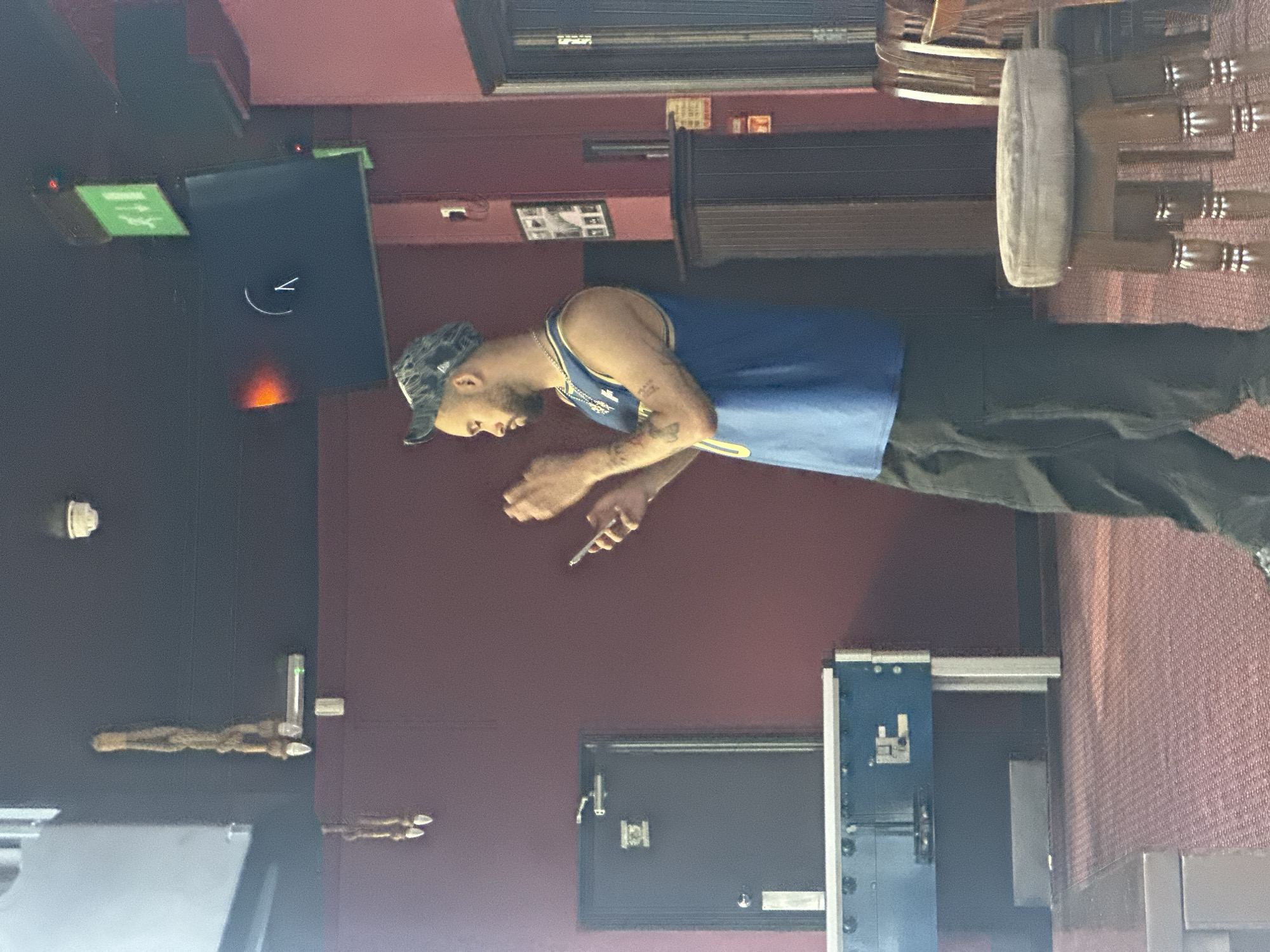 A man wearing a blue and gold tank top, dark pants, and a patterned baseball cap is standing in what appears to be an indoor setting. He is holding an object, possibly a vape pen or similar device, in his right hand. The environment consists of a wall painted in burgundy and dark gray, and a blue locker system is present. The exact location in London is not identifiable from the image.