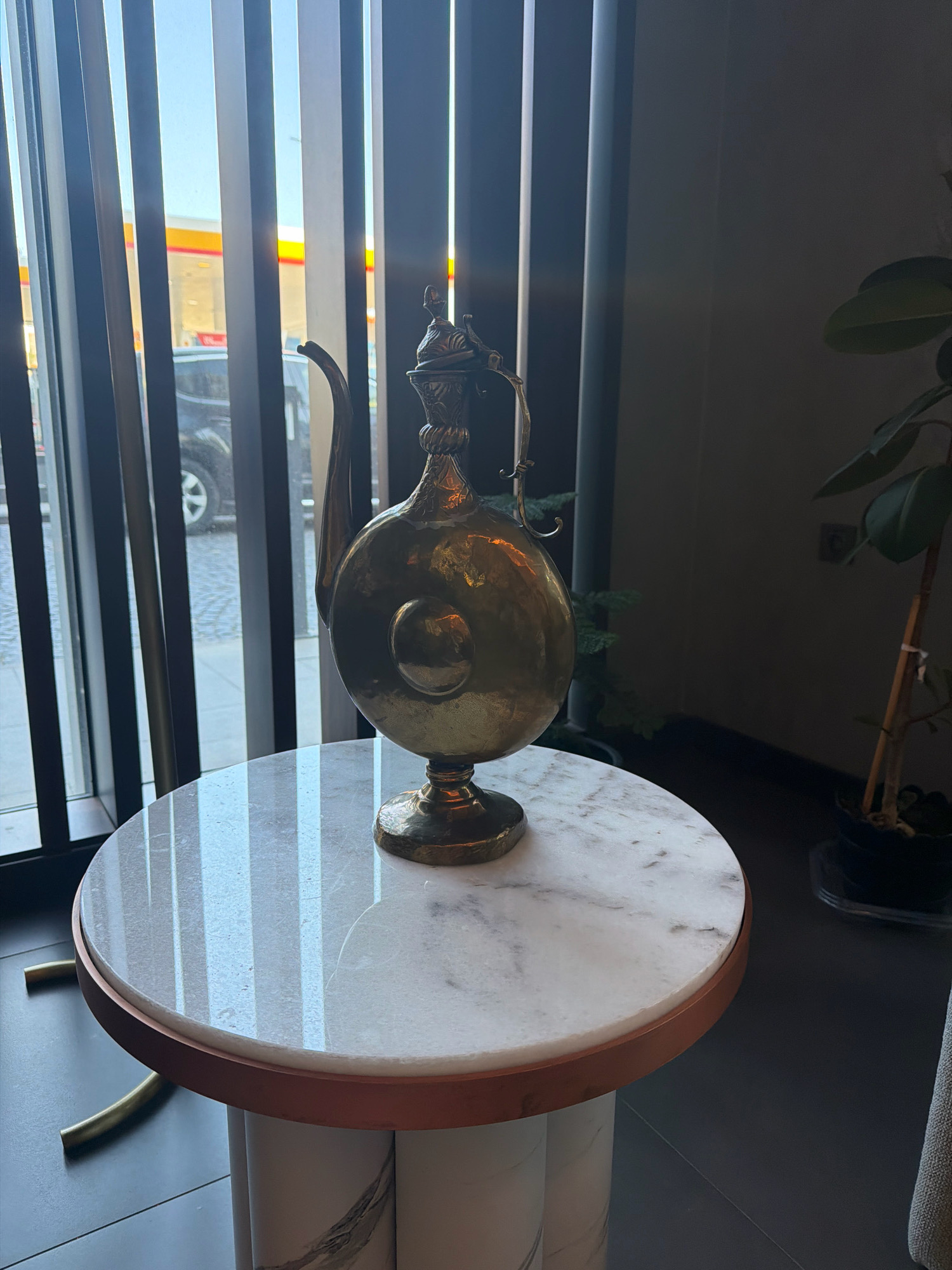 A brass pitcher sits on a marble-topped side table in front of a window with vertical blinds. Outside the window, a street scene is visible, including a car and a yellow and red building which appears to be a gas station. The setting is indoors, likely a cafe or restaurant, given the ambiance. The pitcher is ornately decorated, with a bulbous body and a long spout. The marble table has a copper-colored trim and a cylindrical base with a marbled pattern. A plant is visible to the right of the pitcher. The lighting suggests it is daytime. The location is identified as Bakırköy, Turkey.