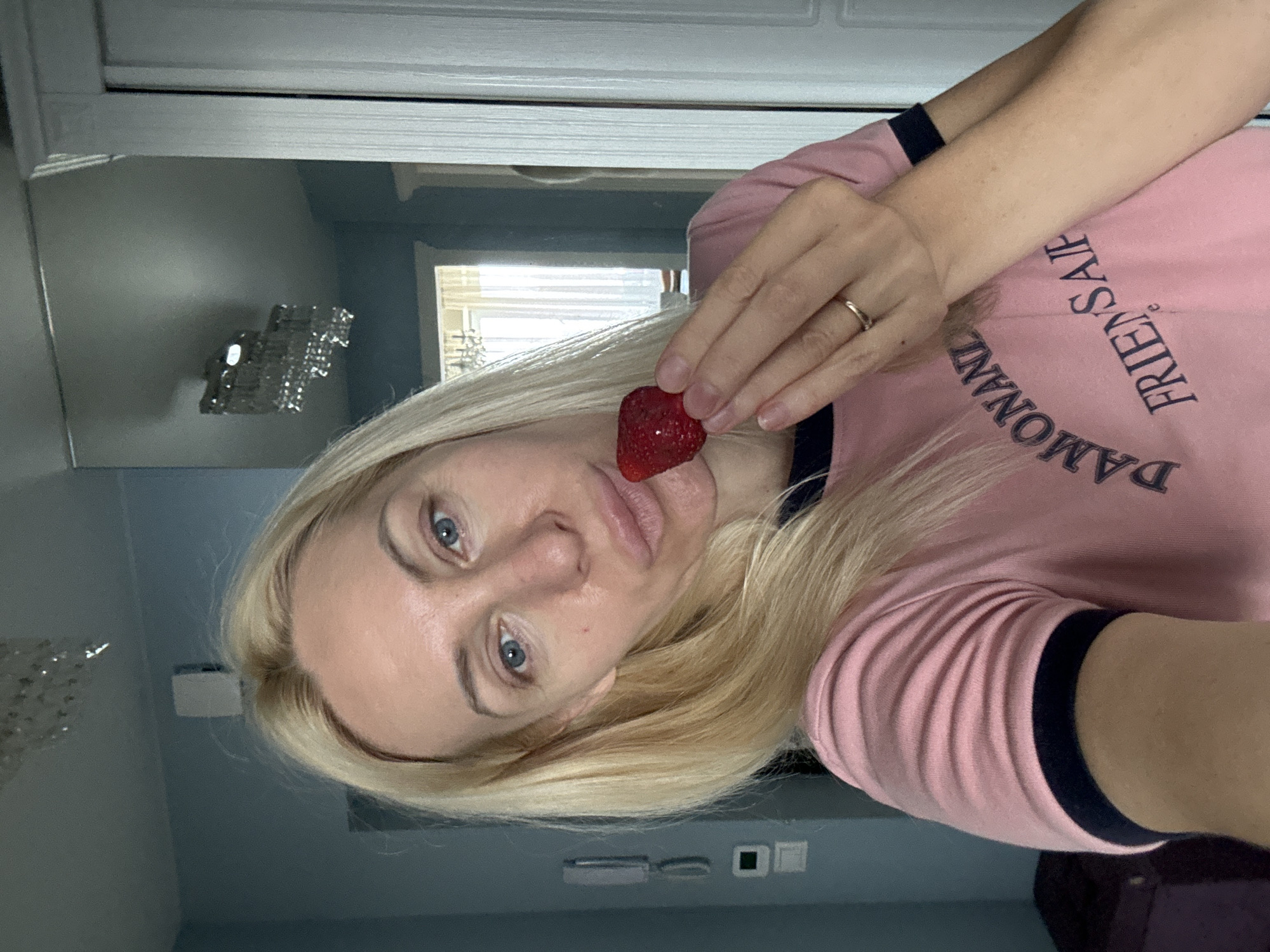 A woman with blonde hair and blue eyes holds a strawberry to her lips in a selfie. She is wearing a pink t-shirt with blue trim and an inscription that reads "DAMONANIZ FRIENDSAIP". Her left hand, adorned with a ring, is visible near her face. The background appears to be an indoor setting, possibly a bedroom or living area, with a light blue wall, a mirror reflecting a chandelier, and a doorway with white trim. The overall lighting suggests daytime.