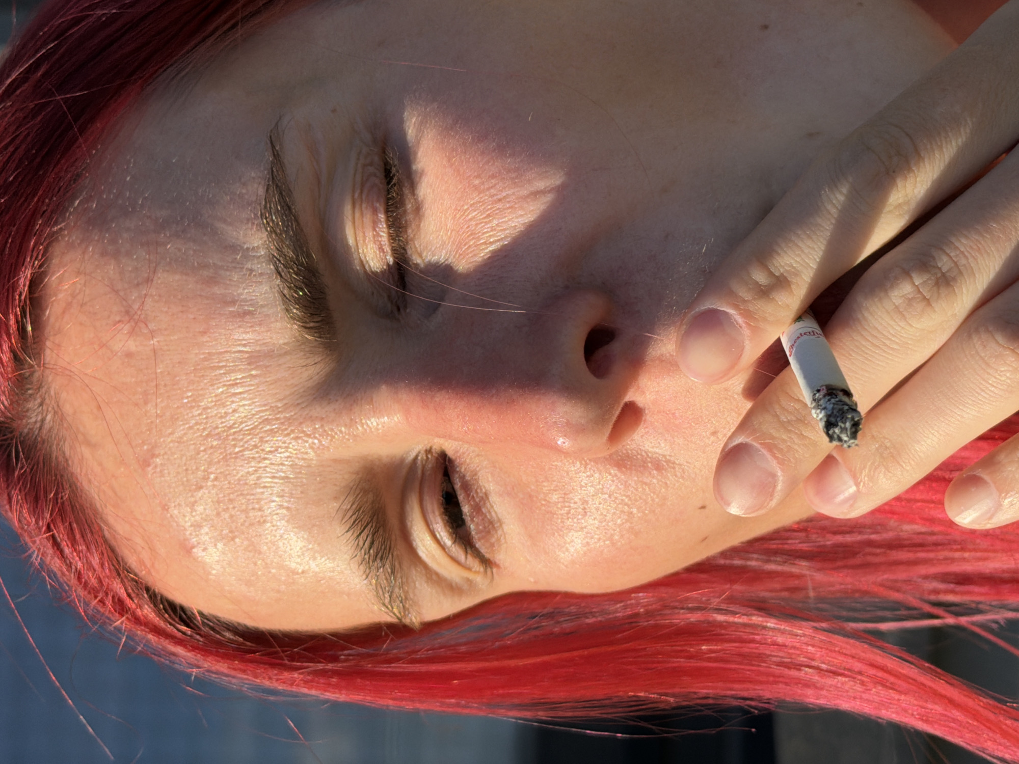 The image is a close-up, low-angle shot of a person with bright red hair, smoking a cigarette. The person's eyes are partially closed, and sunlight illuminates their face, highlighting the texture of their skin. Their fingers are holding the cigarette, which has a lit end. The background is a blurred expanse of blue, suggesting an outdoor environment, possibly near a body of water. The image is a photograph taken in Chaikovsky, Russia.