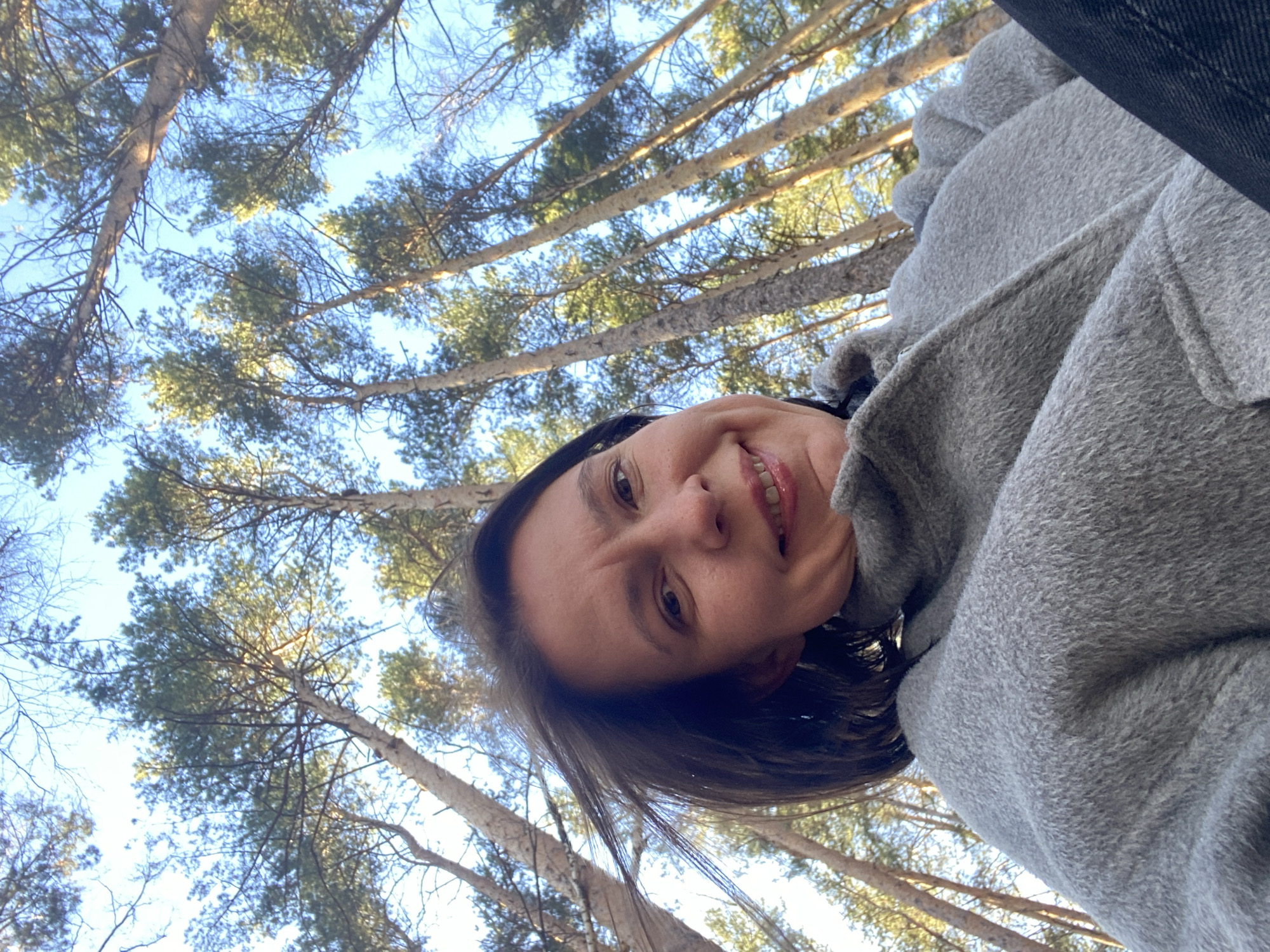 A woman is looking directly into the camera. She is wearing a grey wool coat. The photo is taken from a low angle, looking up into a forest. The trees have thin, bare trunks and green foliage at the top, against a light blue sky. The scene is likely outdoors in a wooded area. No specific location in Chaikovsky, Russia can be determined from the image.