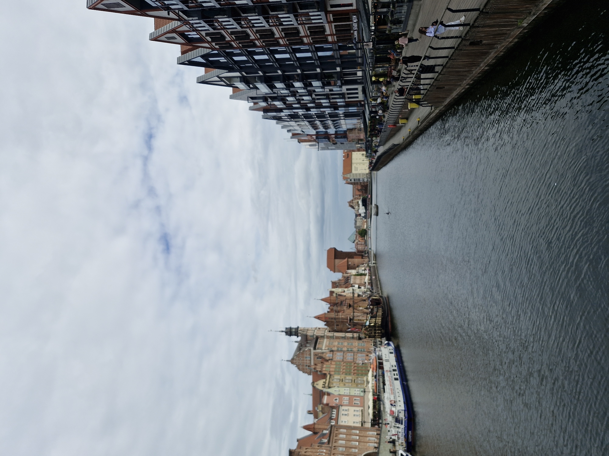 This image depicts a canal-side view of the historic waterfront in Gdańsk, Poland. On the left, a row of colorful, old-fashioned European buildings with steep, tiled roofs line the canal. One of these buildings features a prominent tower. A white and blue passenger boat named "ELŻBIETA" is docked along this side. Further down the canal, a large, dark wooden ship, resembling a pirate ship or historical vessel, is also moored.
On the right side of the canal, a modern building with a dark facade and numerous windows stands in stark contrast to the older architecture. This building has a series of balconies and appears to house shops or restaurants on its ground floor, with awnings extended. A wooden walkway or promenade runs along the edge of the canal in front of this modern building, separated by a railing. Several people are visible on this promenade, some walking and others standing. One woman in the foreground, wearing a white top and pants, is leaning against the railing and looking out at the water. The water itself is dark and rippled, reflecting the overcast sky. The overall atmosphere is that of a well-preserved historical city with modern developments integrated into its waterfront.