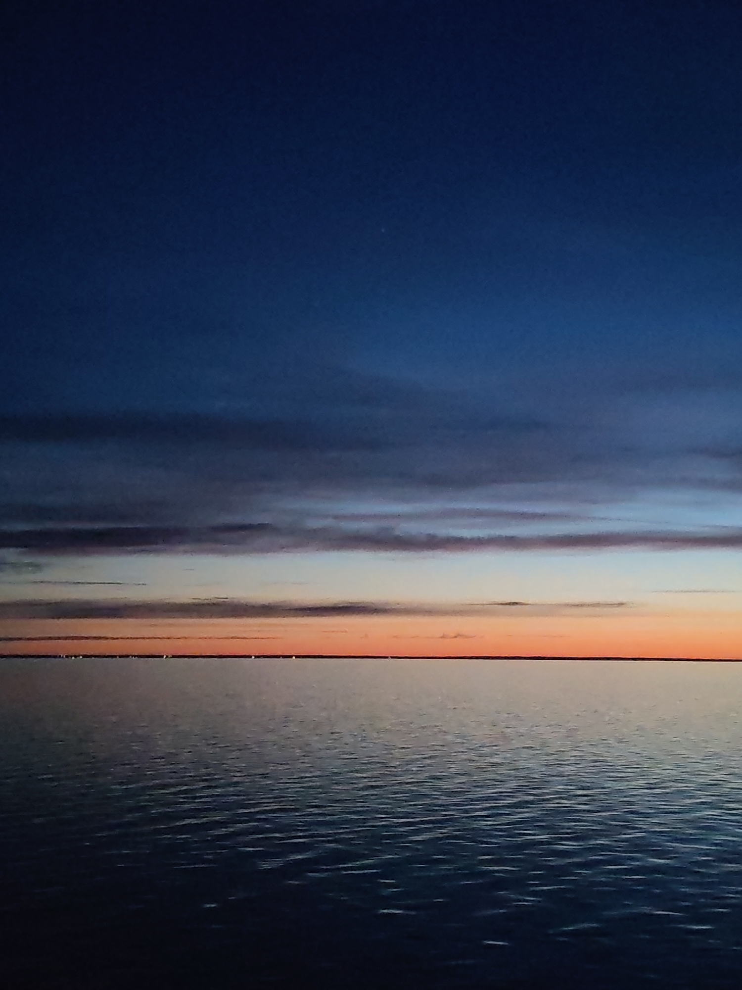The image captures a sunset over what appears to be a large body of water, potentially Lake Beloye, given the reference to Belozersk, Russia. The sky is a gradient of color, transitioning from a dark blue at the top to gray and pink hues in the middle, with a prominent orange band near the horizon. A strip of land is visible on the horizon line, punctuated by what could be city lights or illuminated structures. The water surface is rippled, reflecting the ambient colors of the sky. No people or direct signs of human activity are discernible apart from the distant lights.