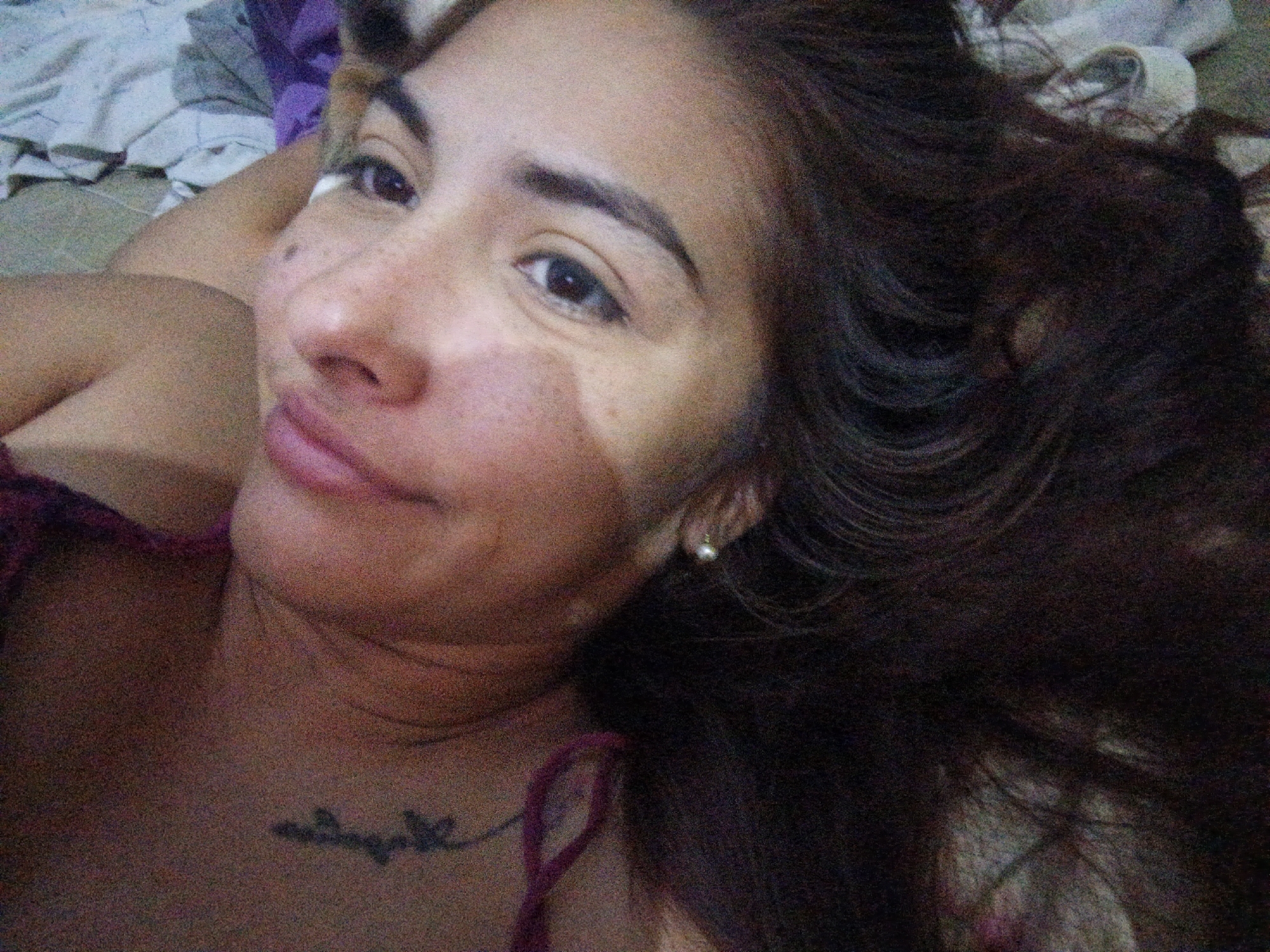 A close-up, angled selfie captures a woman with long, dark brown hair lying on a bed. She is positioned in the foreground, her face dominating the frame. Her skin tone varies, with freckles evident on her nose. A subtle tan line is visible on her forehead and cheek. She has dark eyes, defined eyebrows, and a neutral expression with slightly parted lips. A small pearl earring is visible in her right ear. A tattoo is present on her chest, just below her neck. The woman is wearing a dark-colored top with thin straps. The background consists of a bed covered with wrinkled white and purple bedding. There is no indication of the location being Juan Griego, Venezuela, or any urban setting. The focus is entirely on the woman and her immediate surroundings.