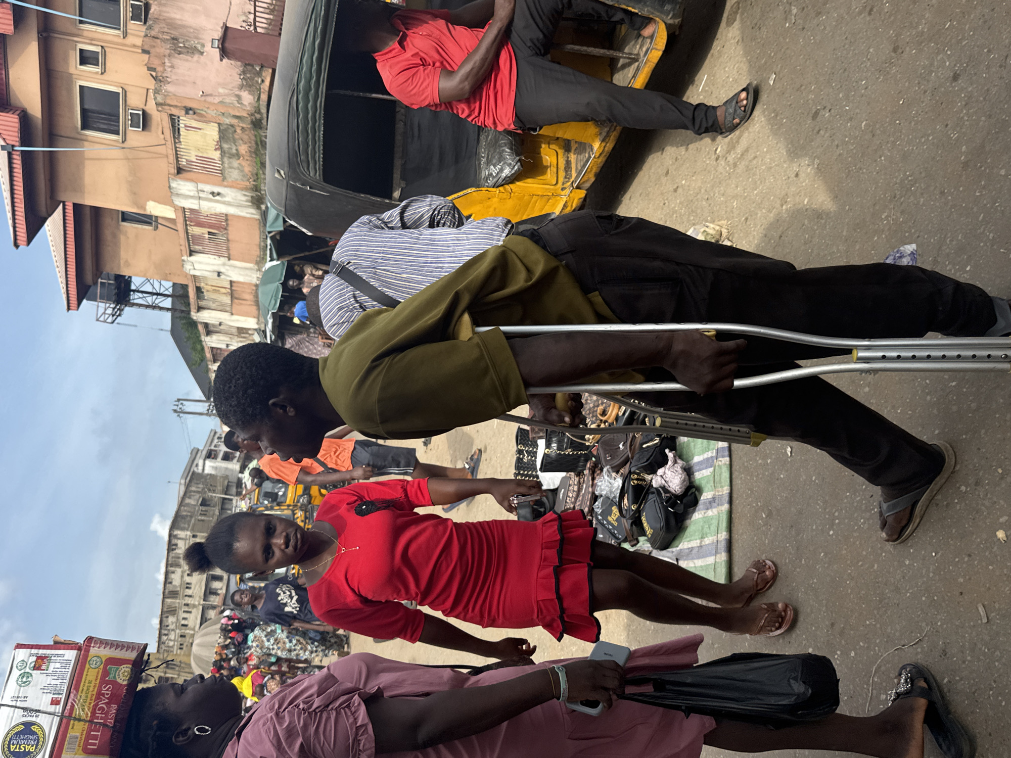 In Nkpor, Nigeria, a busy street scene unfolds under a cloudy sky. A young woman in a vibrant red dress stands near a stall displaying various bags. Beside her, another woman in a pink dress carries a black shopping bag and wears a phone on her wrist. In the foreground, a man on crutches, dressed in olive green and black, walks towards the right. Behind him, a person in a striped shirt is visible, and a yellow auto-rickshaw, a common mode of transport in the area, is parked nearby. A man in a red polo shirt sits on the auto-rickshaw. The environment is bustling with activity, suggesting a market or commercial area, with buildings and stalls lining the street.