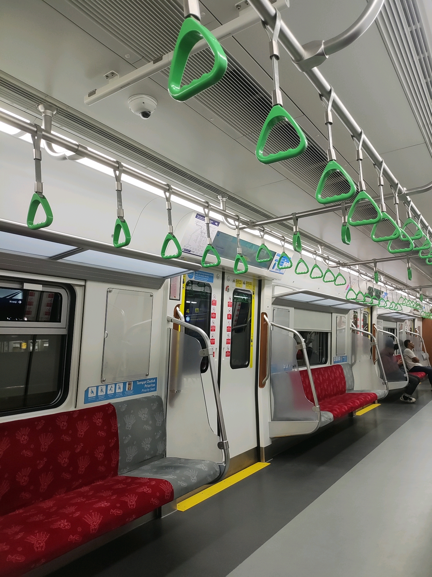 The image shows the interior of a modern train carriage in Bekasi, Indonesia. The carriage is clean and well-lit, with a row of seats on either side and handgrips for standing passengers. The seats are a mix of red and gray, with a patterned fabric. The floor is dark gray with a yellow line indicating the edge of the platform. There are several people in the carriage, some sitting and some standing. One person is visible in the background, sitting on a seat. The train appears to be in motion, as suggested by the blurred exterior visible through the windows. The overall impression is of a functional and efficient public transportation system.