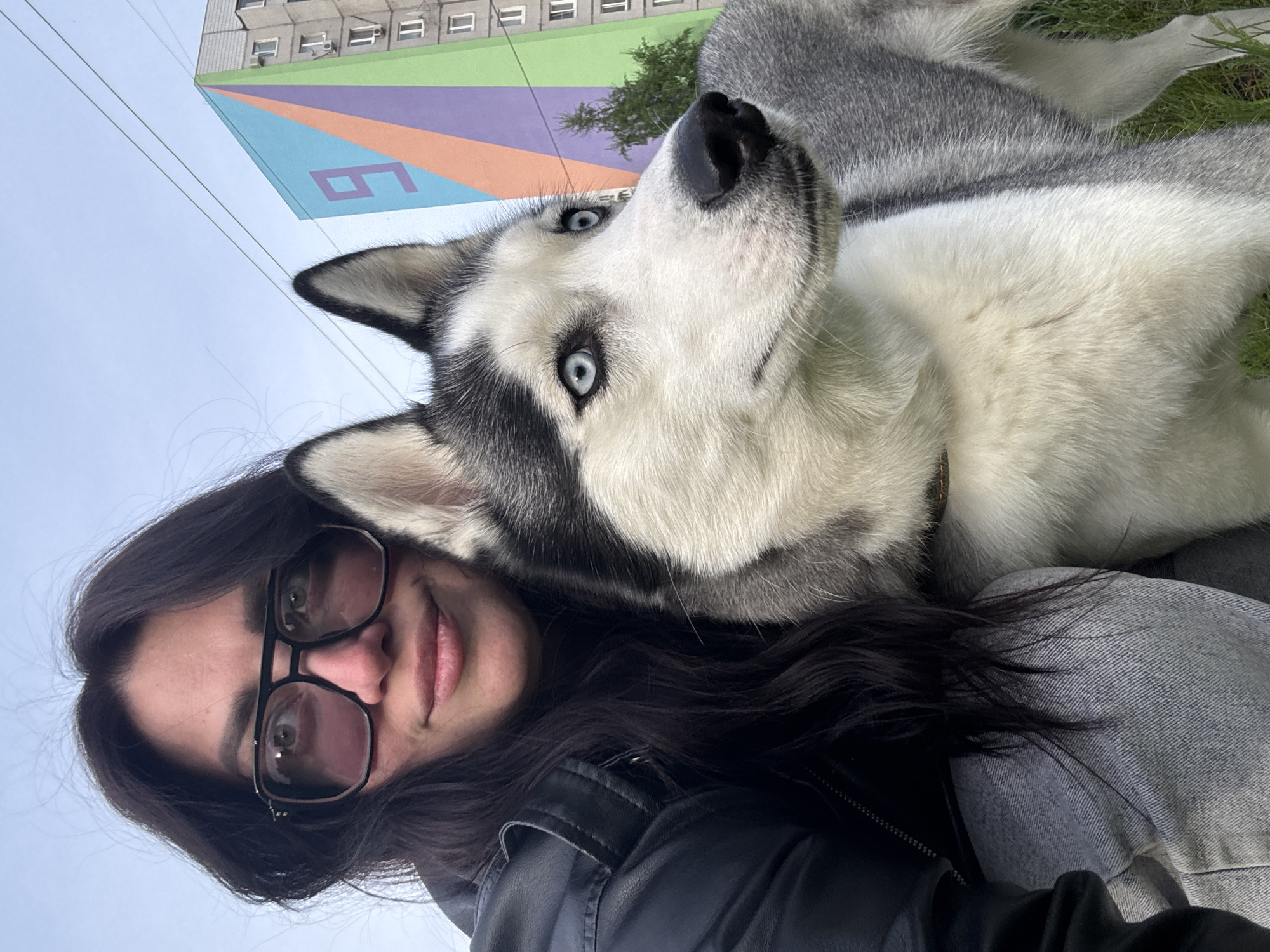 A woman with dark hair and glasses is holding a husky dog in her lap. The dog is looking up at the camera with blue eyes. In the background, there is a building with a mural on the side, and the sky is overcast. The scene is in an urban environment.