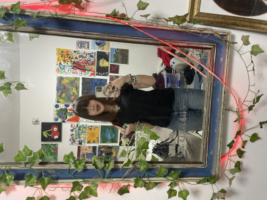 The scene depicts a mirror selfie taken indoors in Raanana, Israel. A young female subject, with shoulder-length dark brown hair and bangs, is reflected in a large, rectangular mirror with a distressed blue and cream-colored wooden frame. She is wearing a black V-neck t-shirt and light blue distressed jeans, and is heavily accessorized with multiple rings on both hands, some featuring green stones, and numerous bracelets on her left wrist. The subject holds a smartphone in her left hand, aimed at the mirror, and has her tongue extended.
The mirror frame is decorated with artificial green ivy vines and an interwoven red LED strip light that emits a distinct red glow. The background, as seen in the reflection, consists of a white wall densely covered with various pieces of artwork, including paintings of different styles and sizes. Notable artworks include a black square painting with two red cherries, a yellow painting depicting a sun-like figure, and a representation of the character Dory. On the floor to the subject's right, visible in the reflection, are a pile of clothing, a small white fan with blue details, and clear plastic storage containers. A portion of another gold-framed object is visible in the bottom right, outside the main mirror's reflection.