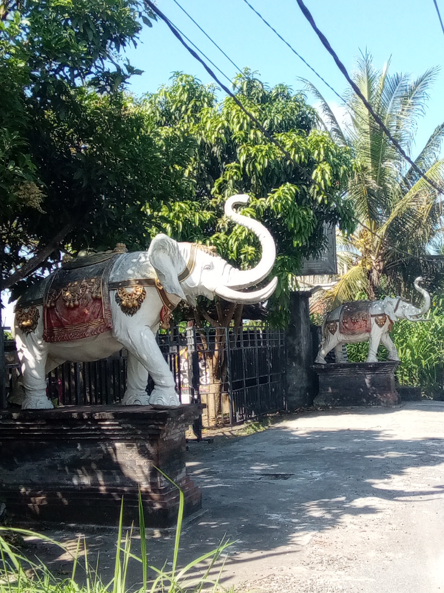 Two decorative elephant statues stand on pedestals in front of a fence. The statue on the left is larger and faces to the right, while the smaller statue on the right faces to the left. Both elephants are white with ornate, colorful coverings on their backs and sides, featuring red, gold, and dark blue details. The larger elephant has intricate gold embellishments and a saddle-like covering. The smaller elephant has a similar, though less detailed, covering. Behind the statues, lush green foliage from trees and palm trees is visible against a clear blue sky. Power lines are strung across the top of the image. The ground in front of the statues is a paved surface, with shadows of leaves and branches cast upon it.