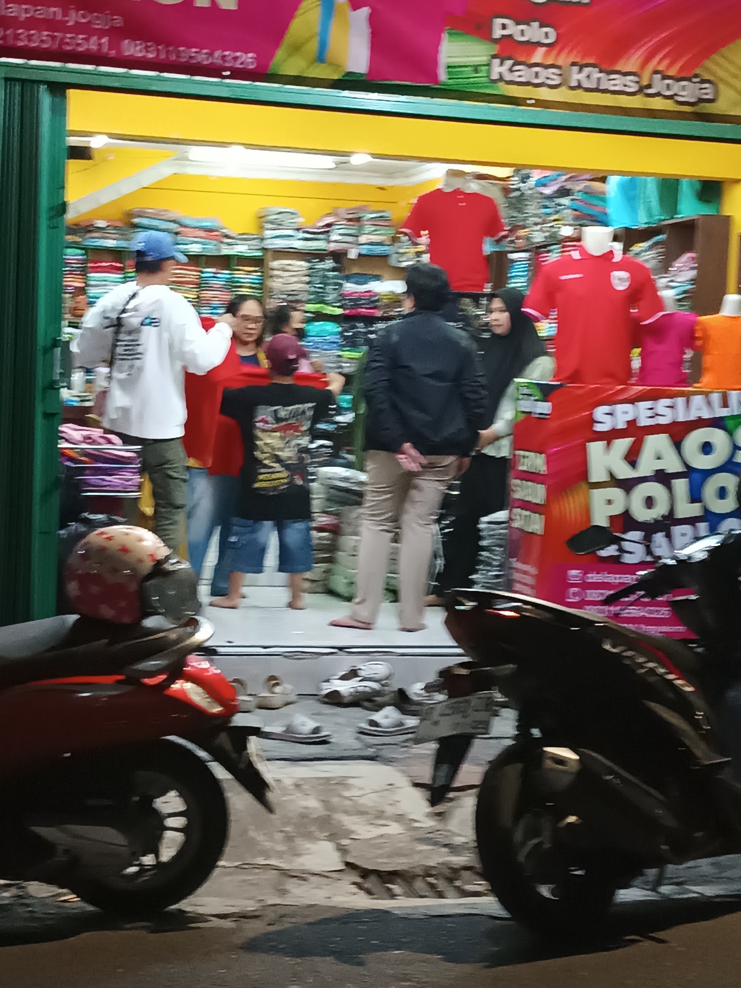 The image depicts a street-level view of a clothing store in Yogyakarta, Indonesia. The store's facade features a green metal gate, and a bright yellow awning. A banner above the entrance advertises "Polo Kaos Khas Jogja," indicating that the store specializes in polo shirts from Yogyakarta.
Inside the store, stacks of folded t-shirts are visible on shelves. Several mannequins are displaying red polo shirts, and one is wearing a pink t-shirt. In the foreground, a person wearing a white long-sleeved shirt and a blue cap is facing away from the camera, holding a bag. Another person in a red shirt is standing next to them. A person in a black jacket and beige pants is also visible inside the store.
Outside the store, two motorcycles are parked in front. The motorcycle on the left is red and has a helmet on its seat. The motorcycle on the right is black. Several pairs of sandals are scattered on the ground in front of the store. The overall scene suggests a busy retail environment on a street in Yogyakarta.