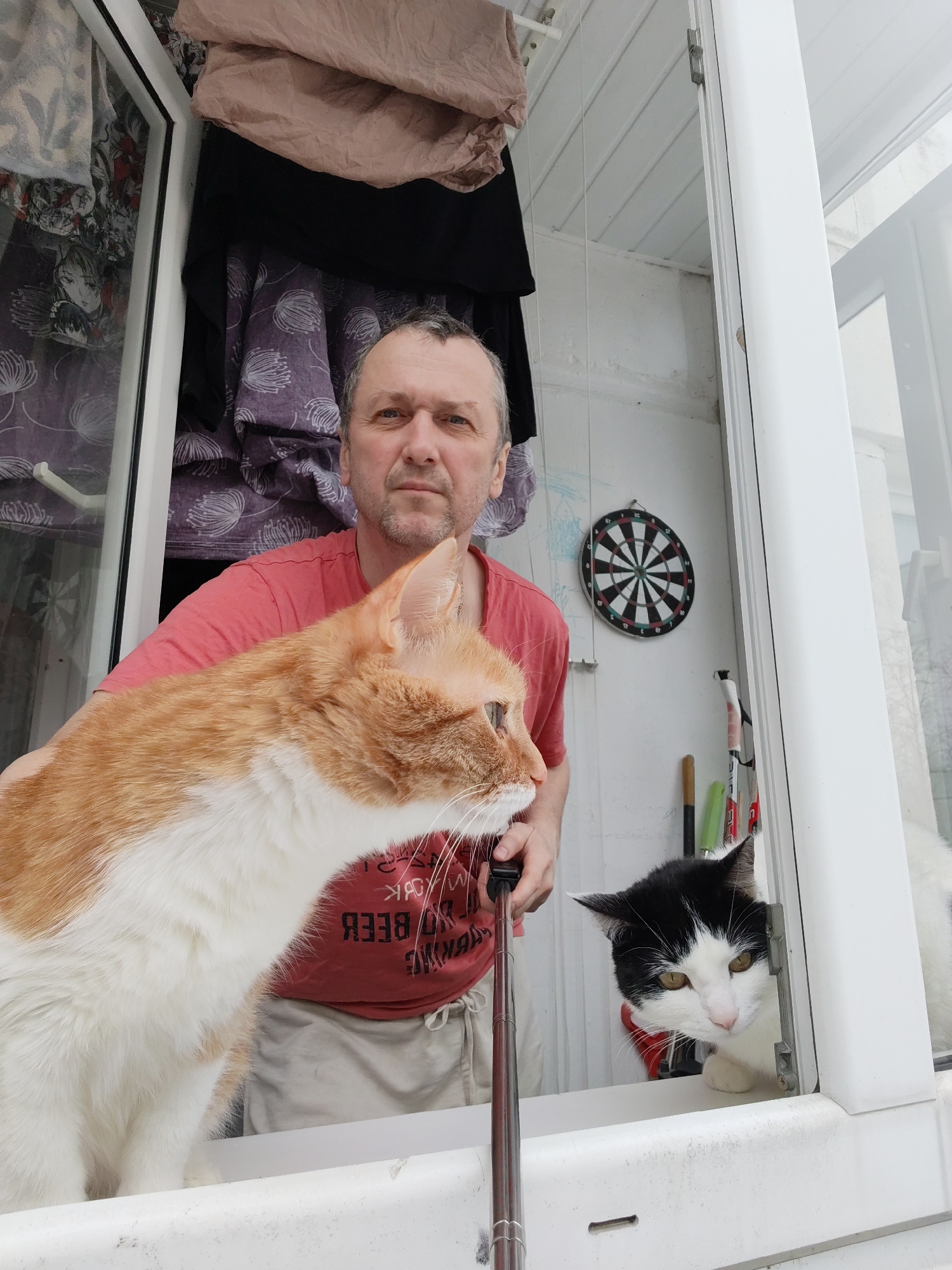 A man is holding a selfie stick, with an orange and white cat in the foreground, while a black and white cat peers through a window in the background. The man is standing on a balcony. In the background, a dartboard is affixed to the wall. Clothing hangs on a rack behind the man. The location is Chaikovsky, Russia.
