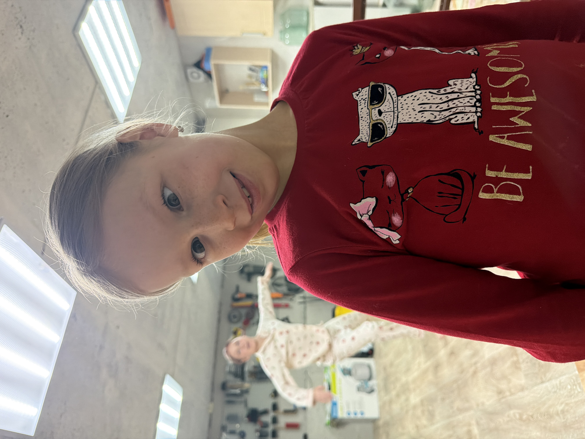The image captures a casual indoor scene, featuring a young girl as the primary subject in the foreground, angled slightly to her left. She has light-colored hair, freckles across her nose and cheeks, and light eyes, looking towards the camera with a gentle smile. She is wearing a red long-sleeved shirt adorned with several cat illustrations. The most prominent cat, in the center of her chest, wears sunglasses.
The shirt also features gold-colored text that clearly reads "BE AWESOME". In the mid-ground, behind the girl and out of focus, another person, an adult woman, is visible. She appears to be wearing light-colored, patterned pajamas.
The background suggests a functional indoor space, possibly a garage, workshop, or utility room. Concrete-like walls are visible, along with shelving units holding various tools and other items, though these details are blurred. The ceiling features several bright, rectangular fluorescent light fixtures, illuminating the room. The overall impression is a candid moment, possibly from a home in Dubovaya, Russia.