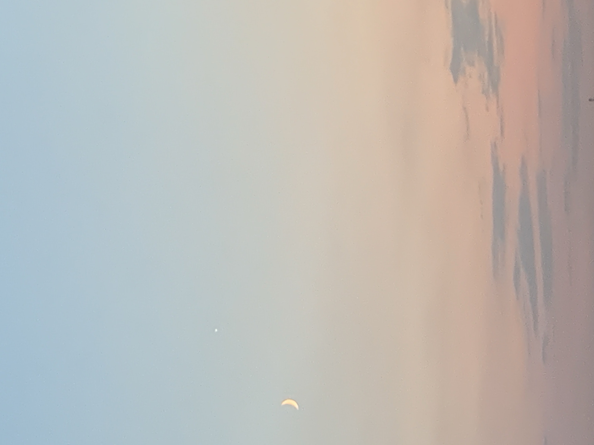 The image depicts a serene evening sky with a gradient of colors, transitioning from light blue at the top to soft pink and peach hues towards the horizon. A crescent moon is visible on the left side of the frame, illuminated by the fading sunlight. A small, bright dot, likely a planet or star, is positioned above and slightly to the right of the moon. wispy clouds are scattered across the lower portion of the sky, adding depth and texture to the scene. The overall impression is one of tranquility and the beauty of twilight. The location of Kolomyia, Ukraine, is not discernible from the image itself.