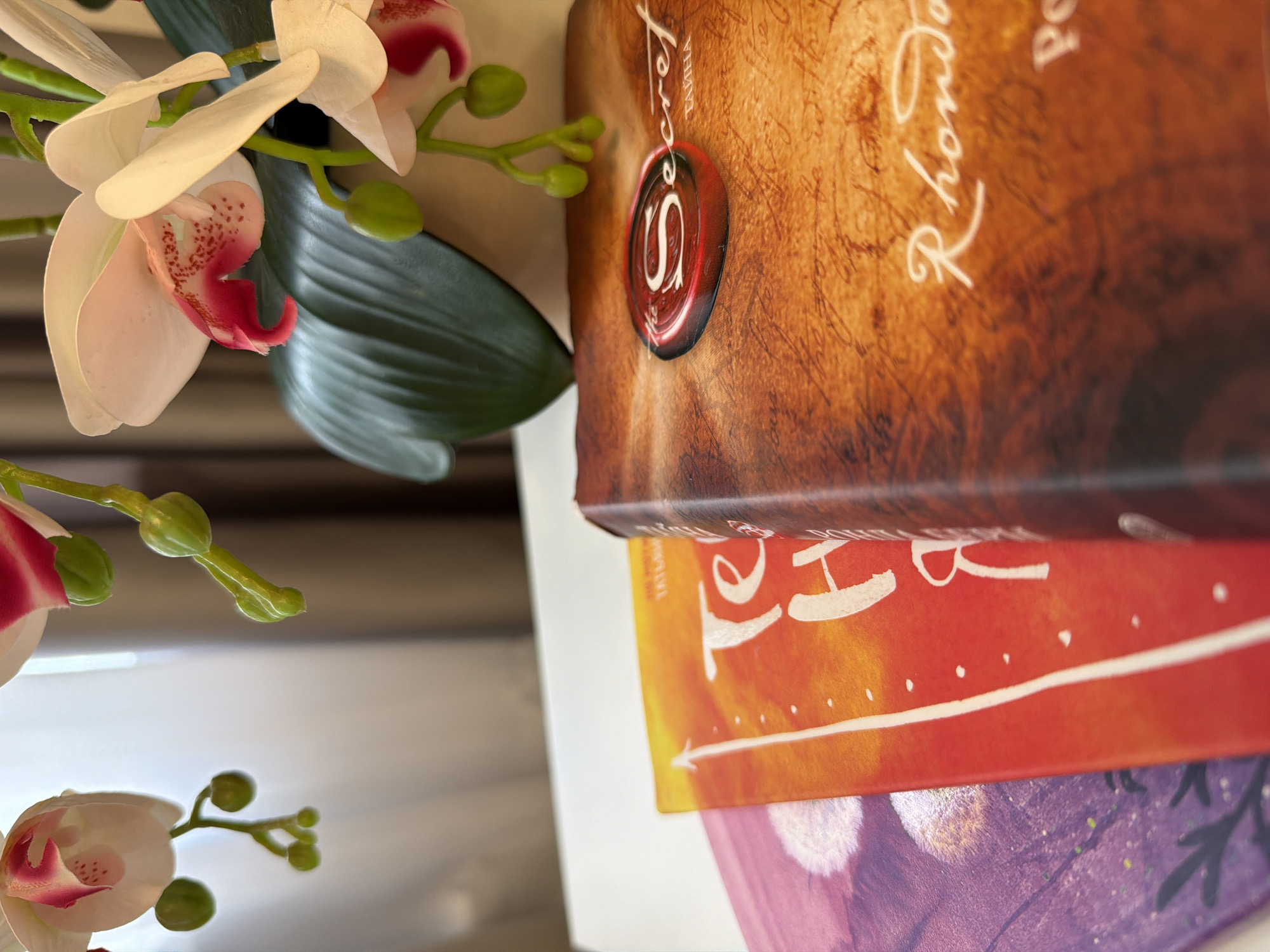 An artificial orchid with white and pink petals and green buds is positioned in the upper right corner of the image. In front of the orchid, three book covers are stacked. The book on the right has a textured brown cover with a red wax seal that reads "The Secret". Below the seal, the name "Rhonda" is visible. The middle book has a bright orange cover with white lettering that appears to say "Te H", and a white line with dots. The leftmost book has a purple cover with white dandelion-like images. The books are on a white surface, and a blurred background of curtains and light is visible behind the orchid. There is no indication of the city of Kazan, Russia.