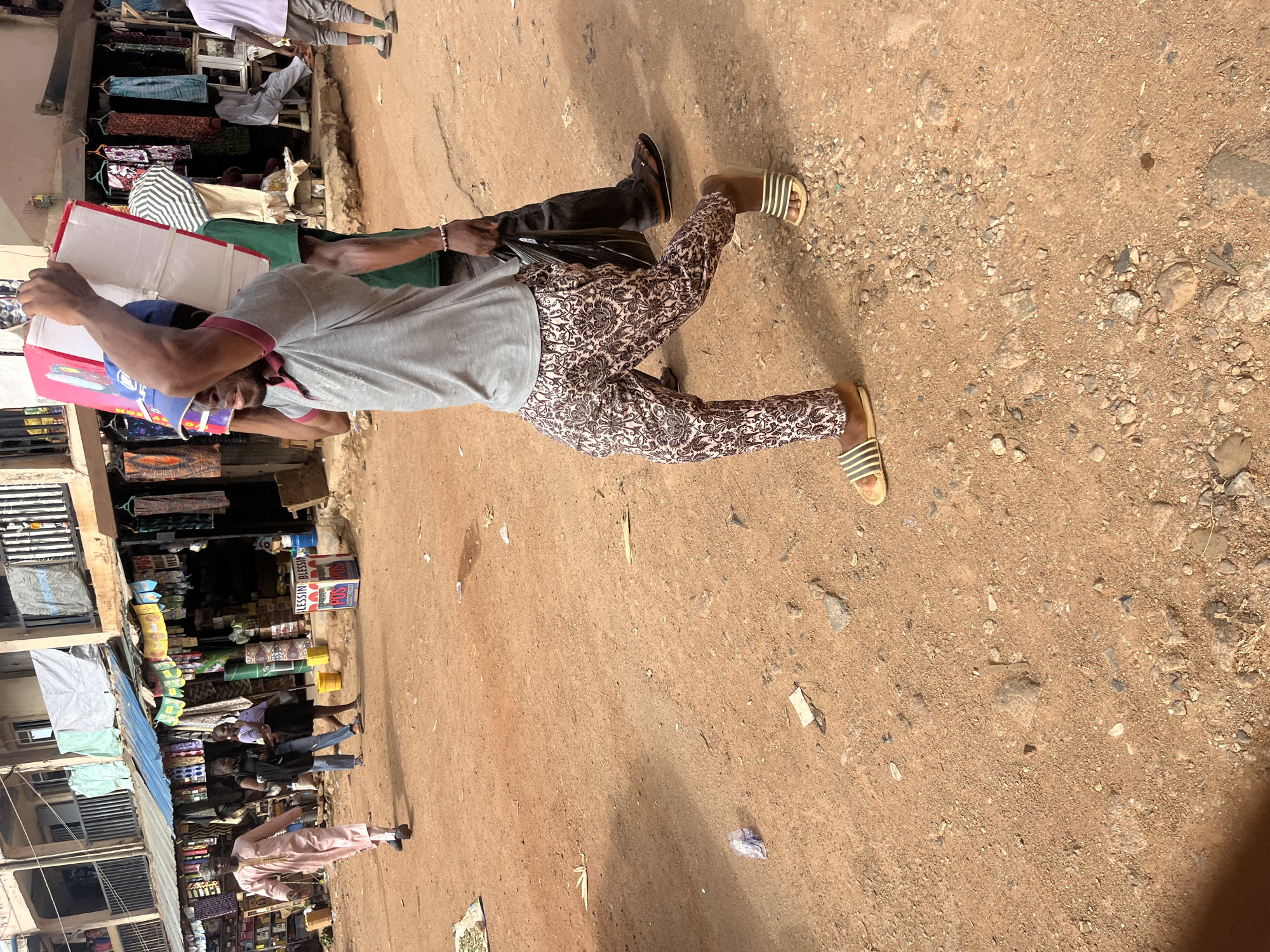 In Jos, Nigeria, a man wearing a blue cap, grey polo shirt, and patterned trousers carries a large, flat box on his shoulders, walking away from the camera. He is accompanied by another person in a green shirt and black pants who walks slightly behind him. In the background, a market scene unfolds with stalls displaying colorful fabrics and goods. Other individuals are visible, including a person in a pink traditional outfit walking towards the market stalls, and two women in the mid-ground. The ground is a dusty, reddish-brown earth mixed with small stones and debris, typical of an unpaved market area. The architecture suggests an urban setting with multi-story buildings and awnings providing shade over the market stalls.