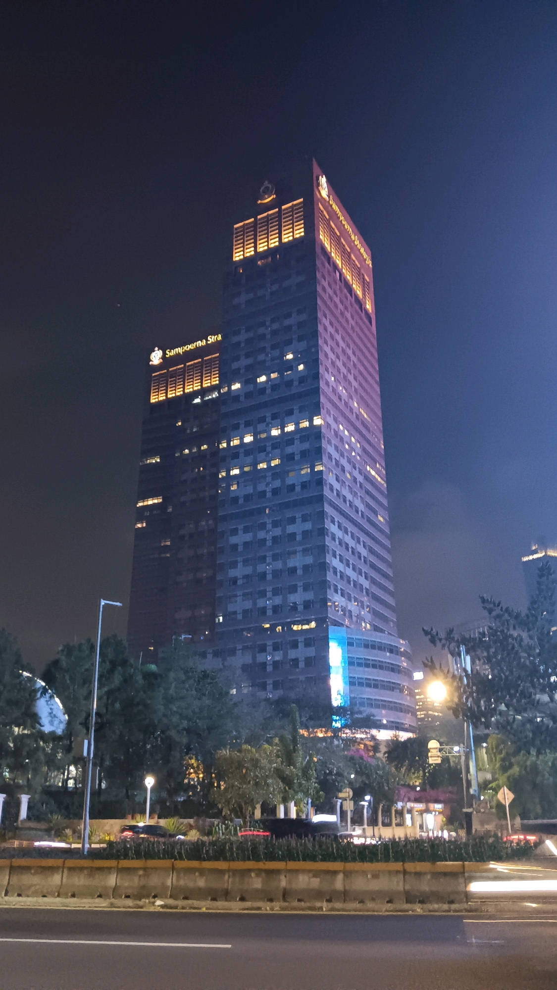 A tall, modern skyscraper with a pinkish hue dominates the frame, its windows illuminated by warm yellow and white lights. The building is labeled with "Sampoerna Stra" in golden lettering near the top. The skyscraper's peak is adorned with illuminated rectangular panels and a circular emblem. To the right, another building with similar lighting is partially visible. In the foreground, a multi-lane road with visible light trails suggests traffic flow. A concrete barrier separates the road from a median strip with trees and streetlights. The sky is a deep, dark blue, indicating nighttime. The overall scene depicts a well-lit urban environment in the city of Central Jakarta, Indonesia.