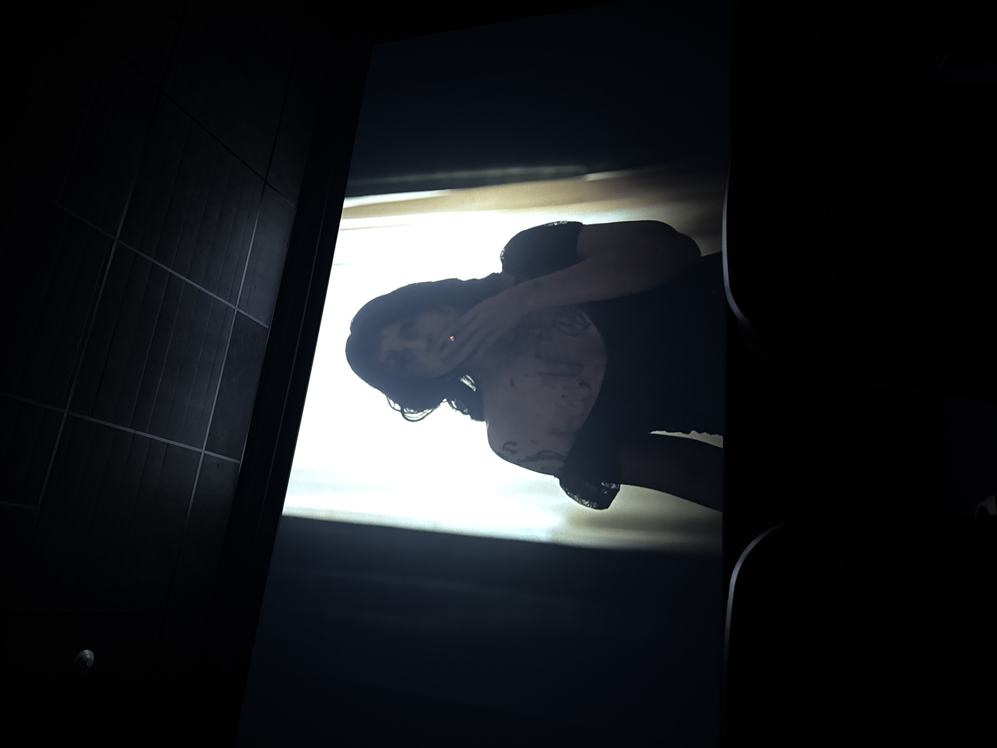 The image shows a woman holding a cigarette, viewed on a screen in a dark room with tile walls. The screen displays a scene with bright light in the background. The woman has dark hair and is wearing a dark top. Tattoos appear on her chest and shoulder. The image does not provide information about a specific location in Puebla, Mexico.