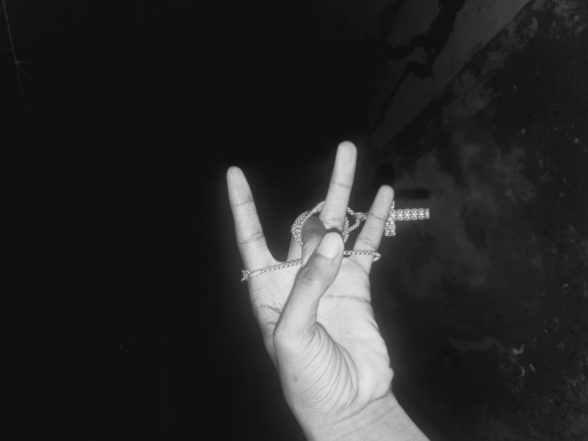 A black and white photograph shows a hand holding a cross necklace. The hand, presumably of a Black person, is positioned with three fingers extended upwards and two fingers folded. The cross necklace is draped across the hand. The background is dark and blurry, but shows a wall with uneven texture. The specific location in Lokoja, Nigeria, is not discernible from the image.