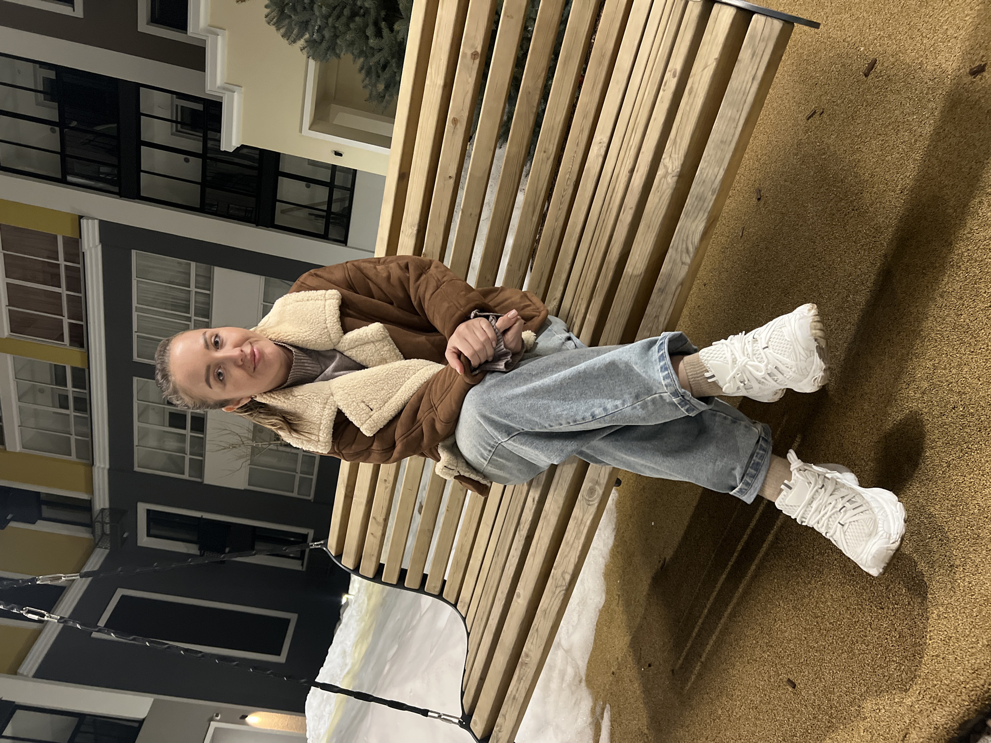 A single female subject is seated on a wooden slatted swing bench in an outdoor urban courtyard at night, located in the city of Unknown, Unknown. The individual, appearing to be in her 20s to 30s, has light skin and light brown hair tied back. She is dressed in a brown faux-suede jacket with cream-colored sherpa lining, a light grey high-neck sweater, light blue cuffed jeans, and white athletic shoes with light-colored socks. Her hands are clasped in her lap, and she maintains direct eye contact with the camera. The swing bench features a dark metal frame and suspension chains. The ground beneath the swing is covered with a tan, granular soft-fall surface, while snow is visible on the ground further in the background and to the left. Multi-story modern apartment buildings with facades of dark grey, light yellow, and white, featuring numerous large windows, dominate the background. Artificial lighting illuminates the scene, and a dark green evergreen shrub is partially visible on the right.