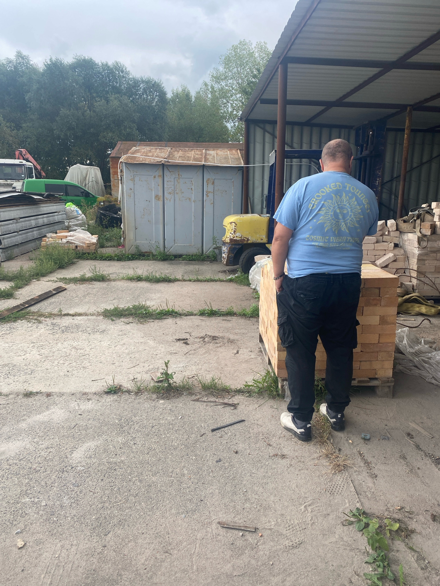 A bald man wearing a light blue t-shirt with a sun graphic and "Crooked Tourist Cosmic Vibes Trips" text, and black cargo pants stands in a dusty outdoor area. He is facing away from the camera, looking towards a building with a corrugated metal roof supported by rusty poles. In front of him is a stack of light brown bricks on a wooden pallet. To his left, there is a yellow forklift and a dilapidated grey shed. In the background, there is a line of trees under a cloudy sky. The ground is a mixture of concrete and dirt with patches of grass and scattered debris.