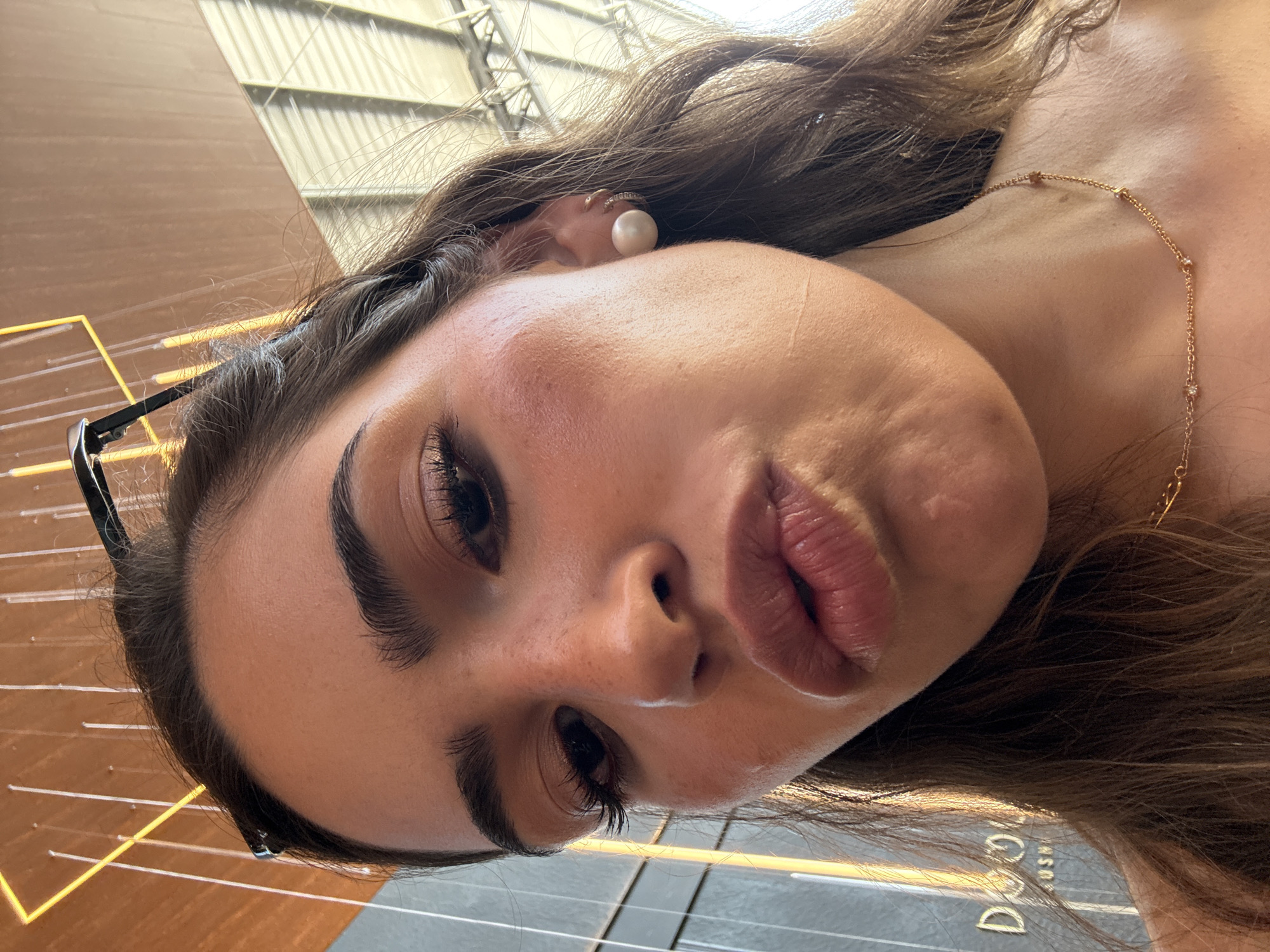 A close-up, tilted image shows a woman's face. She has fair skin, dark eyes, and full lips. Her dark hair is styled with loose waves, and she is wearing a pearl earring and a delicate gold necklace. A pair of eyeglasses are perched on her head. The background suggests an urban environment, with metallic structures and a building sign "DCOOL" visible. The location is San Bernardino Tlaxcalancingo, Mexico.