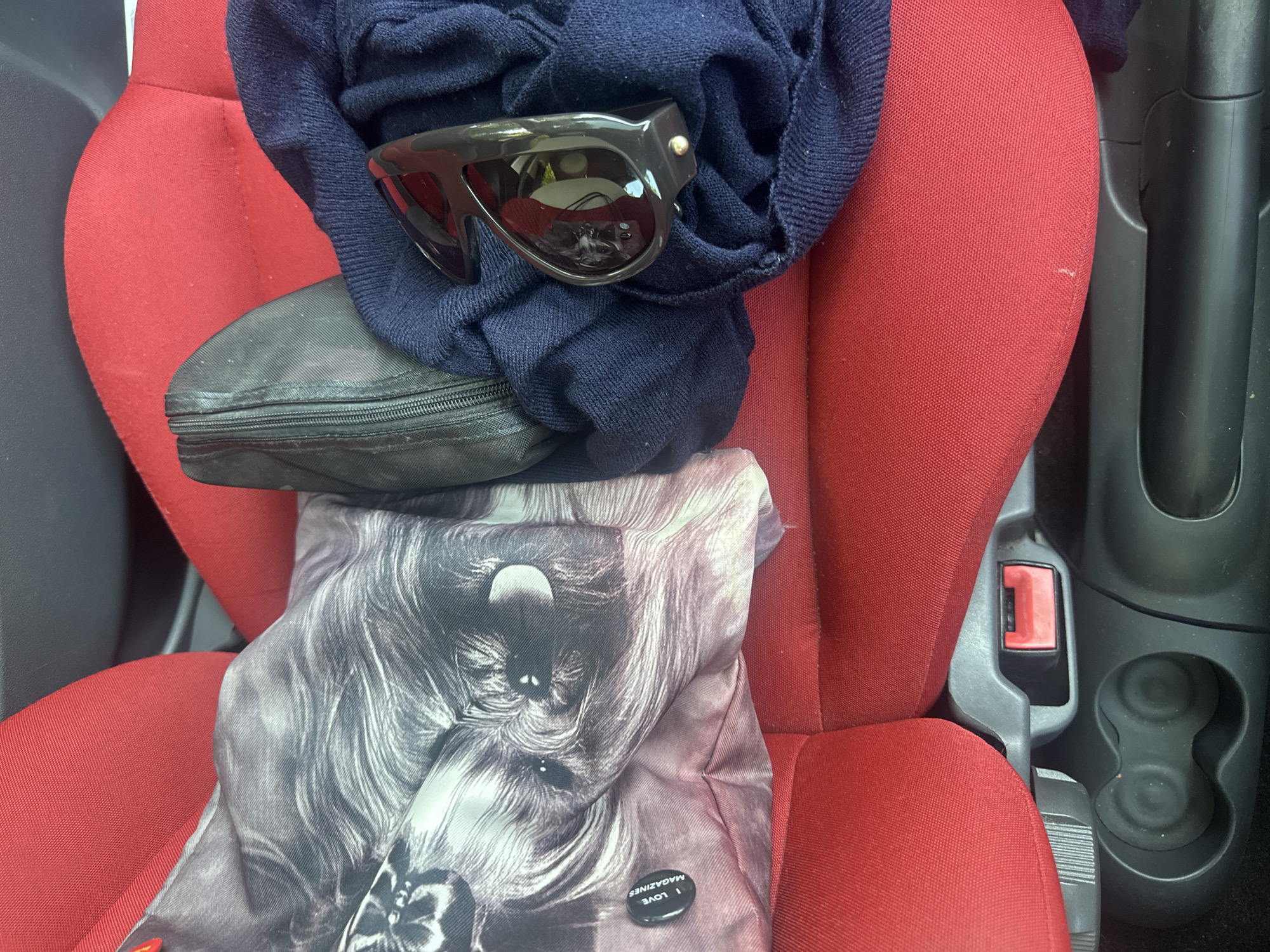 The image captures a collection of personal items resting on a red fabric passenger seat inside a vehicle, likely a car. A dark blue, knitted garment is draped towards the upper left, partially covering a black zippered pouch or case. On top of the blue garment, a pair of black, aviator-style sunglasses with dark lenses is placed.
Below these items, a larger grey and white patterned bag or piece of fabric is visible, featuring a monochrome depiction of a dog's face with shaggy fur. Attached to this bag is a black, circular button or pin. The text "I LOVE MAGAZINES" is clearly visible in white capital letters on the button, curving along its edge.
To the right of the red seat, a black plastic car console is visible, featuring two circular cup holders and a red-buttoned seatbelt buckle receiver. The lighting suggests it is daytime. No human subjects are present, and no activity is occurring beyond the placement of these objects. The setting is clearly the interior of a vehicle, in Waverley, United Kingdom.