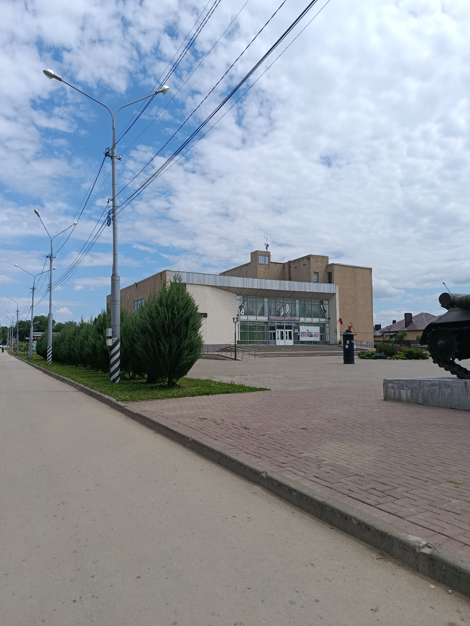 The image shows a street scene in the city of Marx, Russia, with a modern building on the right. The building has a large glass facade and appears to be a public institution, possibly a cultural center. In front of the building, there is a paved plaza with a monument that resembles a tank. A row of manicured evergreen bushes lines the walkway leading to the building. A street with power lines and streetlights runs along the left side of the image, with an asphalt road and a sidewalk. The sky is partly cloudy. A person is visible in the distance walking on the sidewalk. The scene appears to be a public area, possibly a town square or a park.