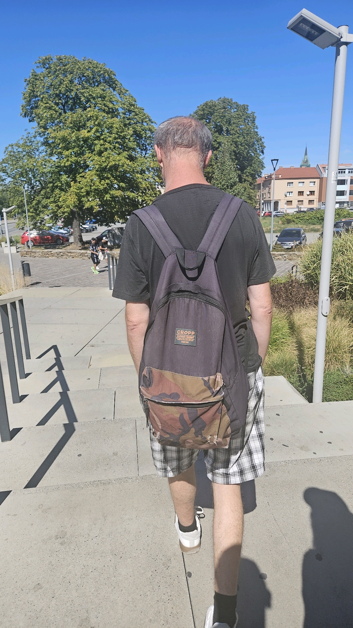 A man wearing a black t-shirt and plaid shorts walks up concrete stairs, carrying a dark backpack with a camouflage bottom. The backpack has a "CROPP" logo. He is wearing white sneakers with black socks. In the background, there are trees, parked cars, and buildings, suggesting an urban environment. The scene appears to be outdoors during daylight, with a clear blue sky. The location is identified as Prešov, Slovakia.