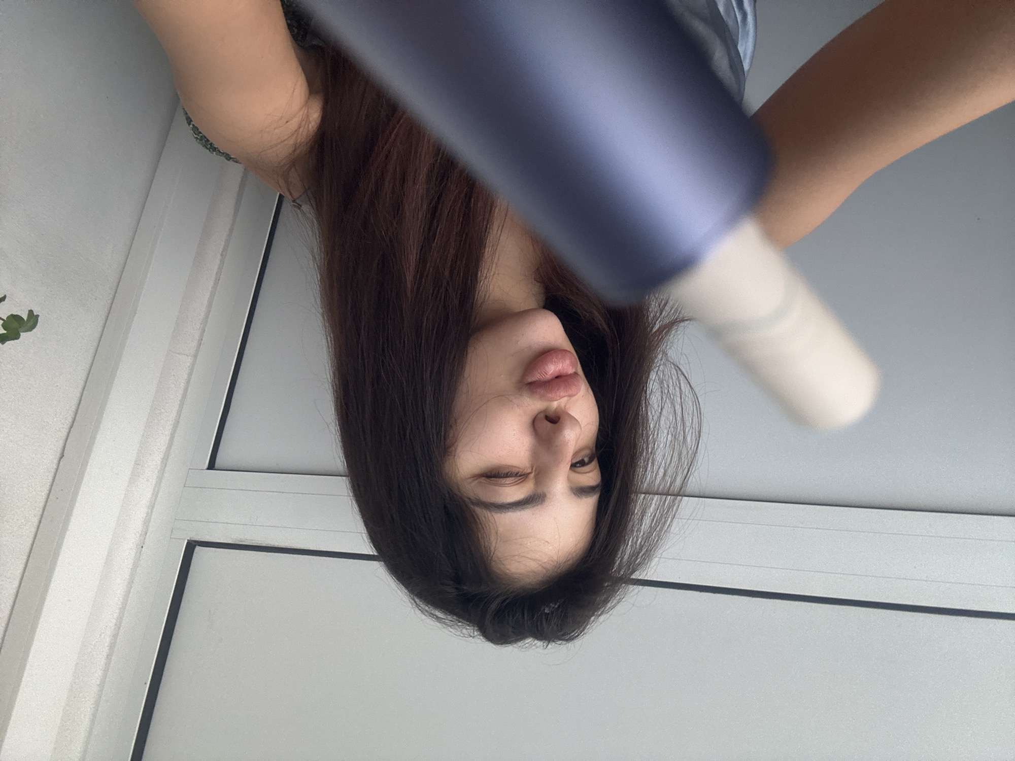 An upside-down, close-up shot features a young person with long, dark brown hair. The subject's head is inverted, facing downwards from the top of the frame. One eye is closed in a wink, while the other is open, gazing slightly upwards. The lips are lightly puckered in a playful or humorous expression.
A large, blurry, dark blue/purple object, possibly a hair dryer or similar styling tool, extends diagonally across the upper part of the image, obscuring part of the subject's forehead and hair. Its end transitions to a lighter, almost white or cream color. The subject's light-skinned arm and shoulder are visible on the right side of the frame, wearing what appears to be a light-colored top.
The background suggests an indoor setting with white walls and a window or door frame composed of white panels and black outlines. A hint of green foliage from a plant is visible in the bottom-left corner. The lighting is bright, indicating an indoor environment, likely during the day. No visible text is present in the image. The overall mood conveyed is lighthearted and candid due to the subject's playful expression and the unusual perspective. The image is contextually located in Plovdiv, Bulgaria.