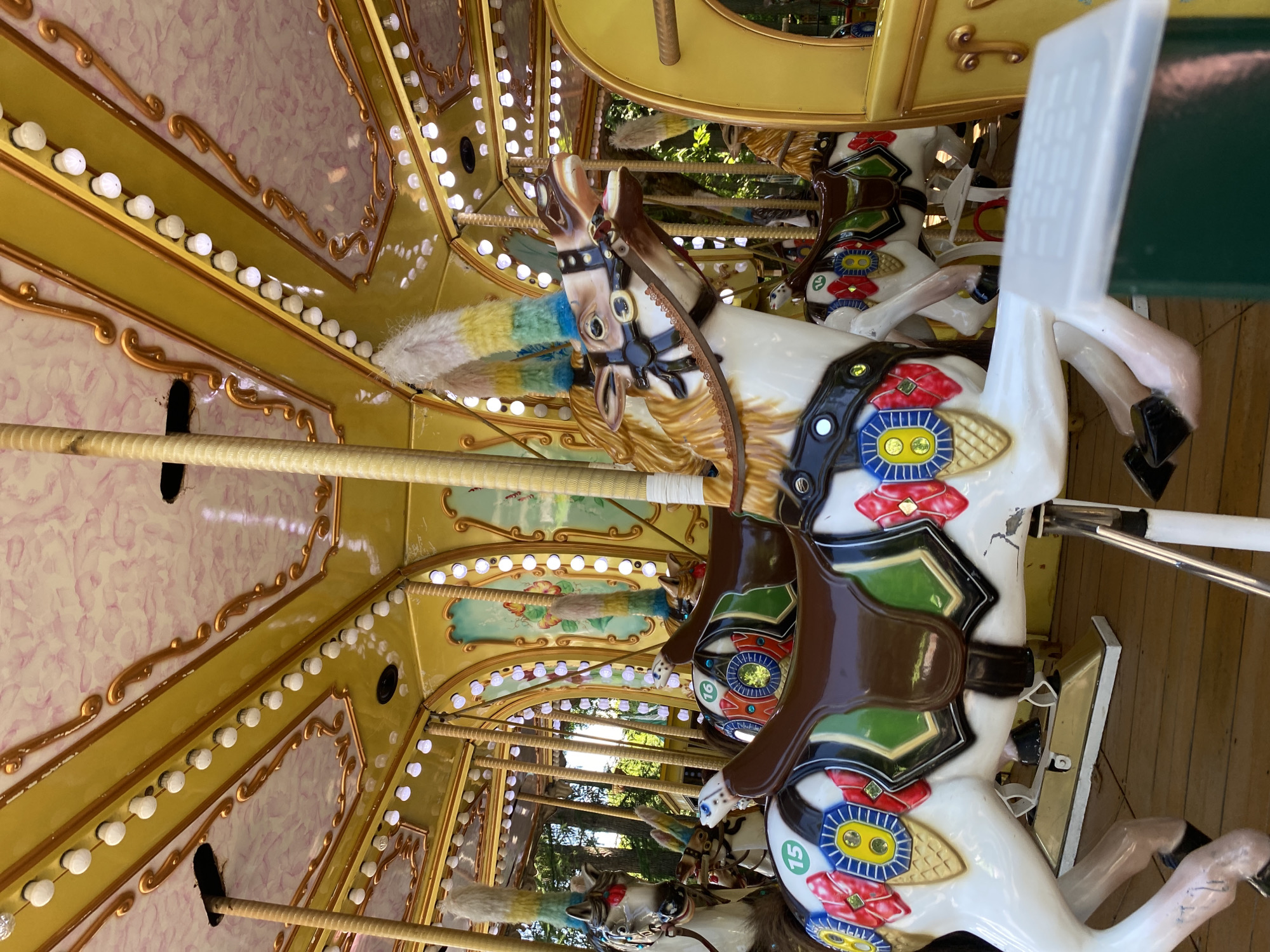 A carousel with brightly painted horses is depicted. The carousel has a yellow and gold decorative ceiling with numerous small white lights. Several horses, adorned with colorful saddles and plumes on their heads, are visible. One horse in the foreground is white with a brown saddle and a decorative bridle. It has the number "15" on its flank. Another horse behind it has the number "16". The carousel is situated on a wooden floor. In the background, trees and foliage are visible, suggesting an outdoor location, likely a park or amusement area. The overall scene is festive and colorful. The image does not provide any information about the city of Perm, Russia.