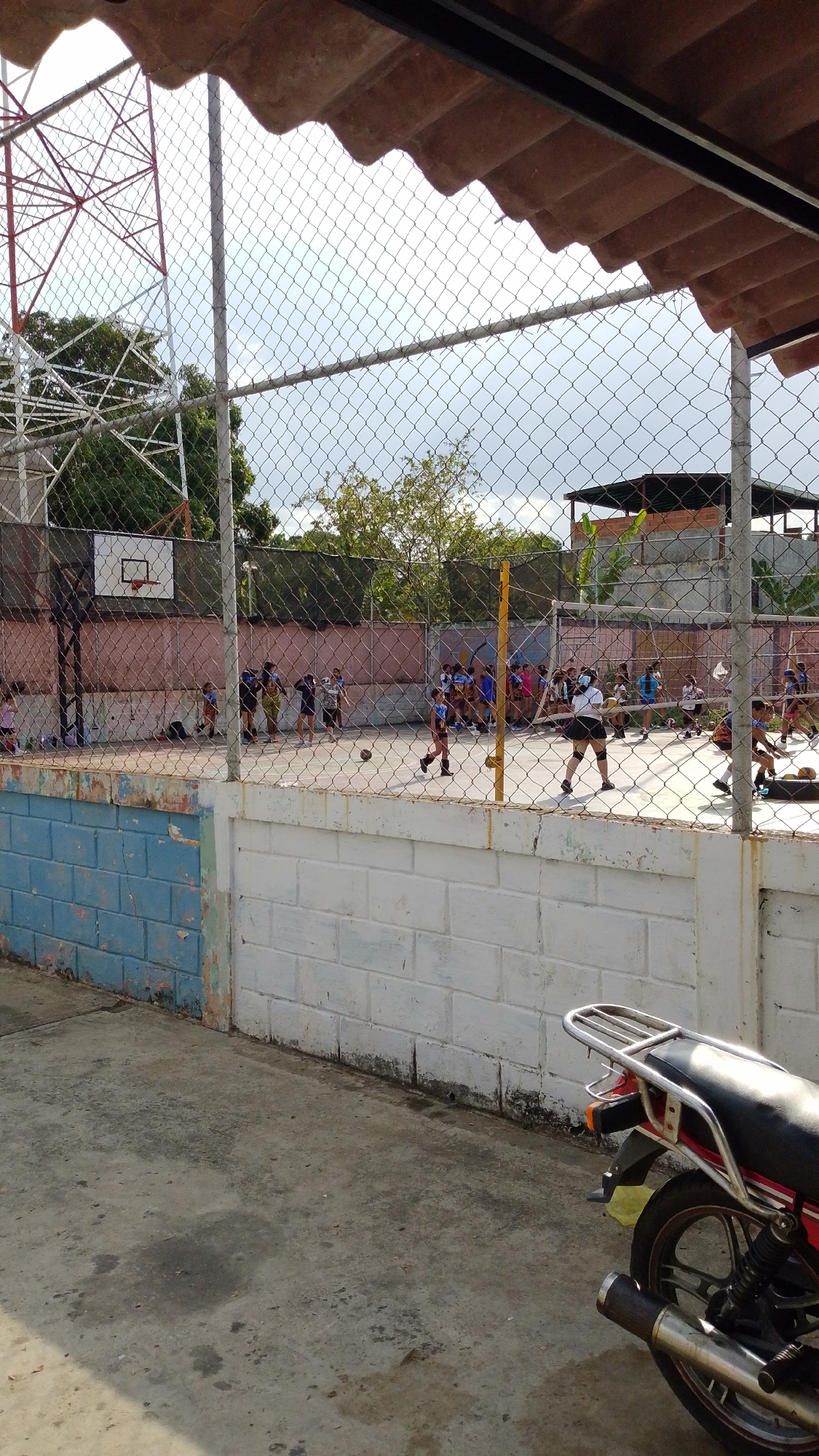 The image shows a chain-link fence separating the viewer from a sports court in Tinaquillo, Venezuela. A group of people is playing a game on the court. There is a basketball hoop on the left side of the court and a volleyball net in the center. A white brick wall runs along the front of the court, partially painted blue. A motorcycle is parked in the foreground to the right. Behind the court, there is a partially constructed brick building with a flat roof. A tall communication tower stands to the left of the court.