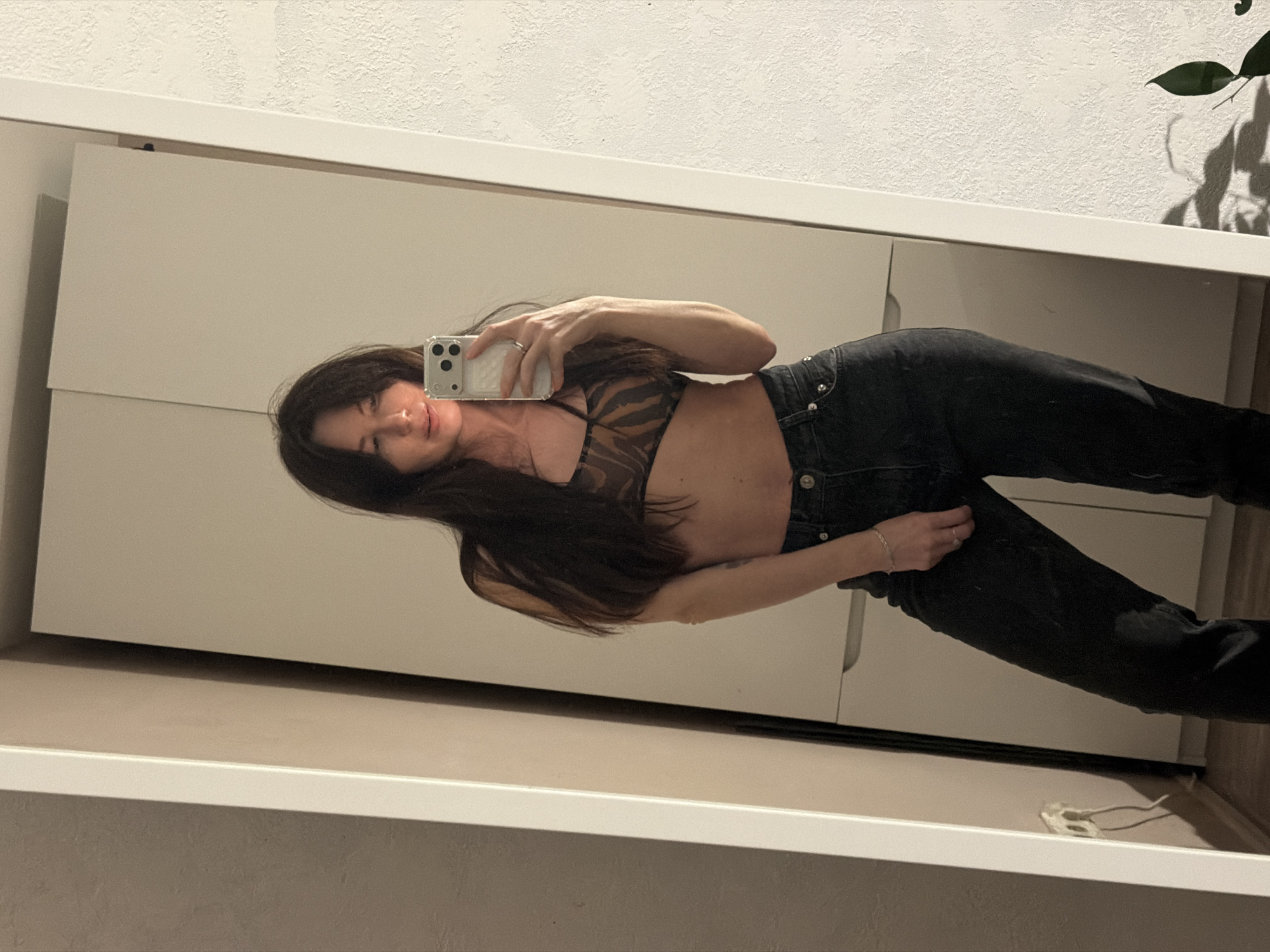 A woman is taking a selfie in front of a mirror. She is wearing a patterned top and black jeans. The background shows a white wall and a closet. The photo does not provide enough information to determine if the location is in Perm, Russia.