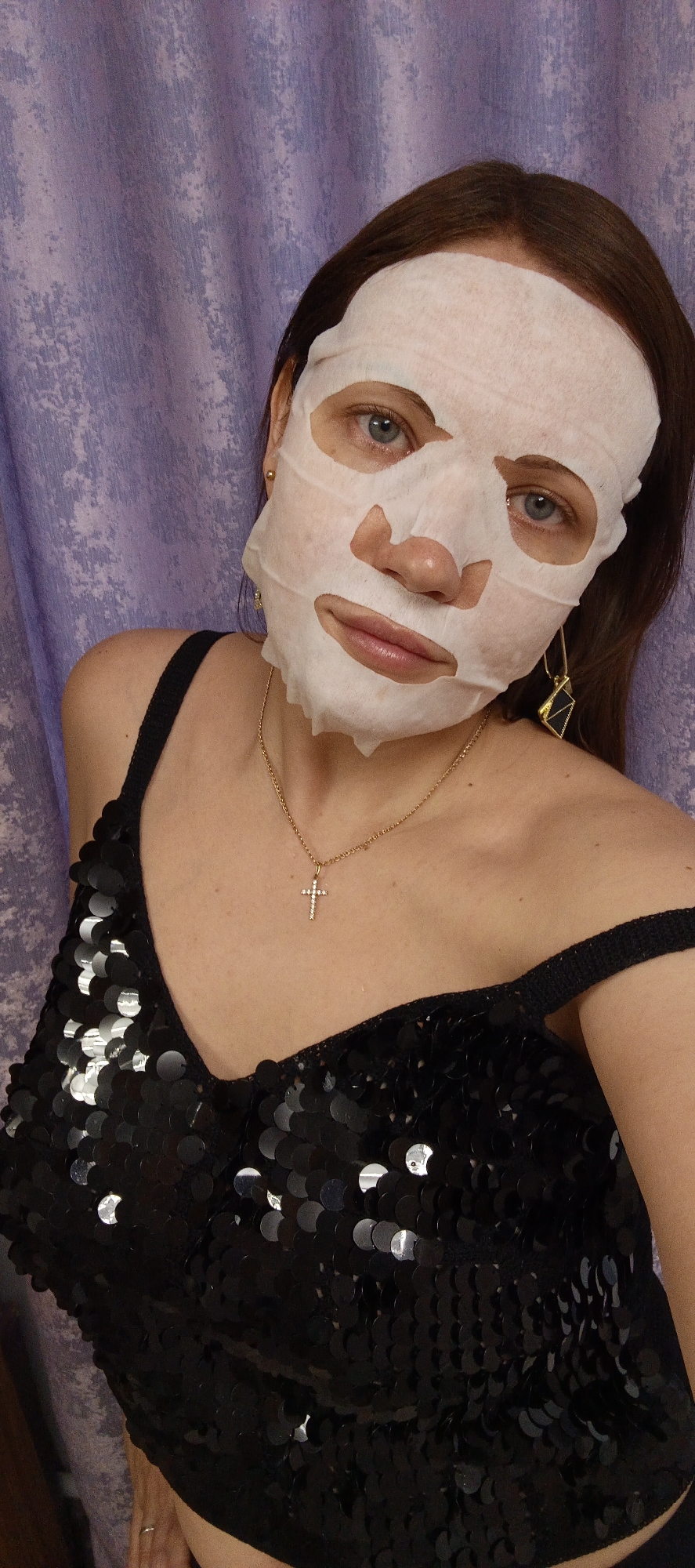 A woman is posing indoors against a light purple patterned background. She wears a white sheet mask on her face. The woman has brown hair and blue eyes. She is wearing a black sequined top with thin straps, a gold chain with a cross pendant, and gold dangle earrings. The location is potentially Chaikovsky, Russia.