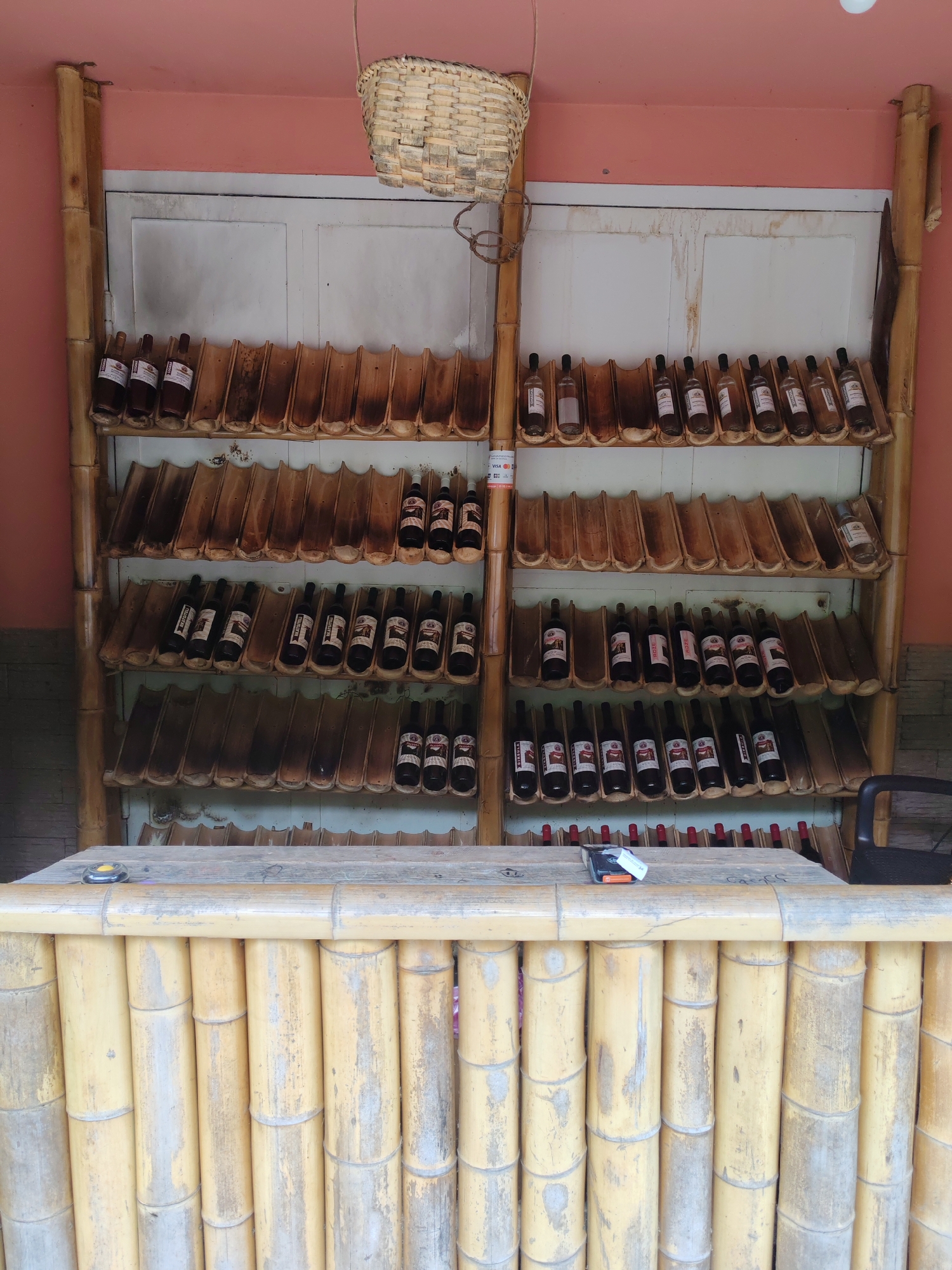 The image depicts a display of bottled beverages, likely wine or spirits, arranged on tiered shelves made of bamboo. The shelves themselves are designed with carved bamboo segments that cradle the bottles. The backdrop appears to be a white, paneled wall, possibly a door or cabinet. Above the shelves, a woven basket hangs suspended. The entire structure is framed by vertical bamboo poles. In the foreground, a bar constructed from vertical bamboo stalks is visible, with a wooden countertop on which a small black device with an orange button rests.
The lighting suggests it is daytime, and the setting appears to be an indoor or covered outdoor area, given the wall and the lack of natural light indicative of open sky. The overall aesthetic is rustic and natural, utilizing bamboo extensively in both the display and the bar. The bottles are varied in color and labeling, some appearing dark, others lighter, suggesting different types of beverages. There is no discernible activity or people in the image.
Some faint text is visible on labels on the bottles, but it is too indistinct to read. A small label with "VISA" is visible on the left side of the middle shelf. The location context of Pirveli Maisi, Georgia, suggests this might be a local winery, distillery, or a bar with a focus on regional products.